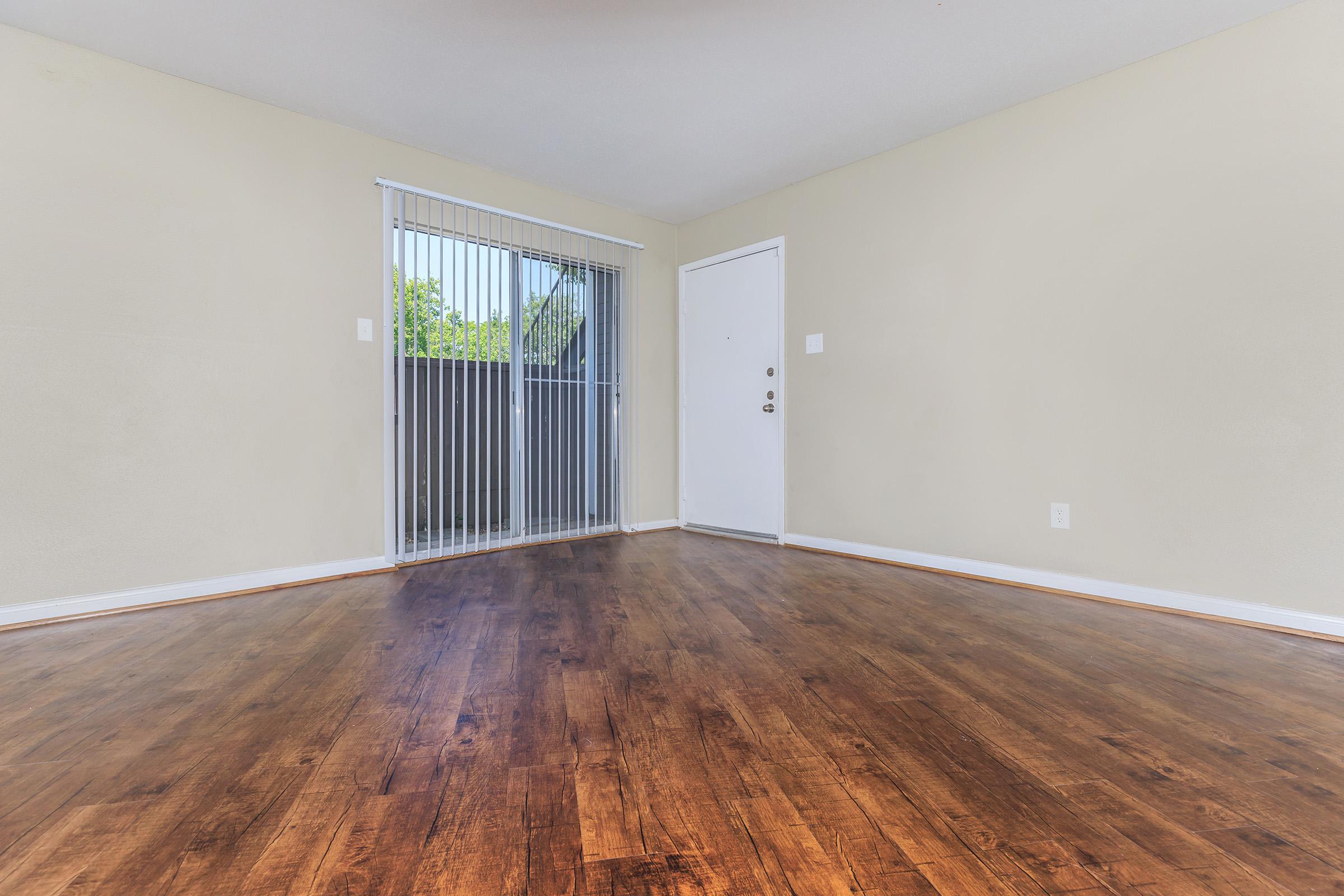 A well-lit empty room featuring hardwood flooring and a large window with blinds. The walls are painted a light color, and there is a white door on one side. The space appears clean and ready for furnishing, with a straightforward layout that highlights its openness.