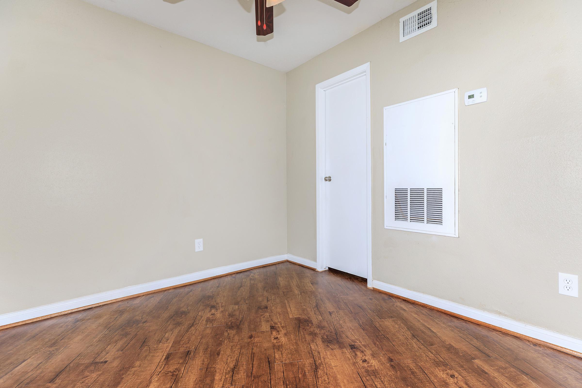 A sparsely furnished room featuring light beige walls, a ceiling fan, and hardwood flooring. There is a closed white door on one side and a wall-mounted vent near the corner. The room has an overall clean and simple appearance, with minimal decorations or furniture.