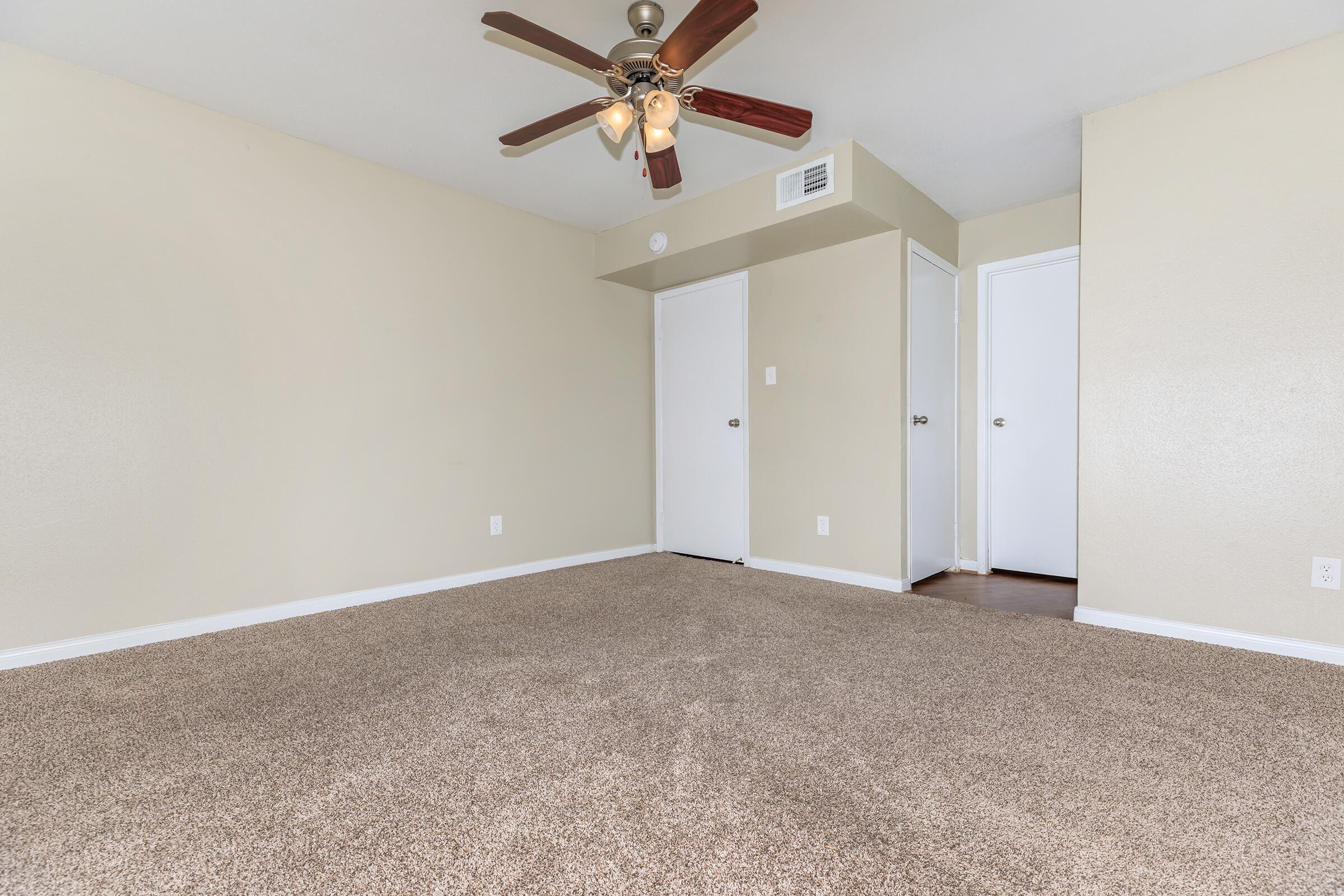 A spacious, empty room with light beige walls and a plush carpet. A ceiling fan with wooden blades hangs from the ceiling. There are two closed doors on one side, one leading to a closet and the other to a different space, contributing to the room's simple and welcoming atmosphere.