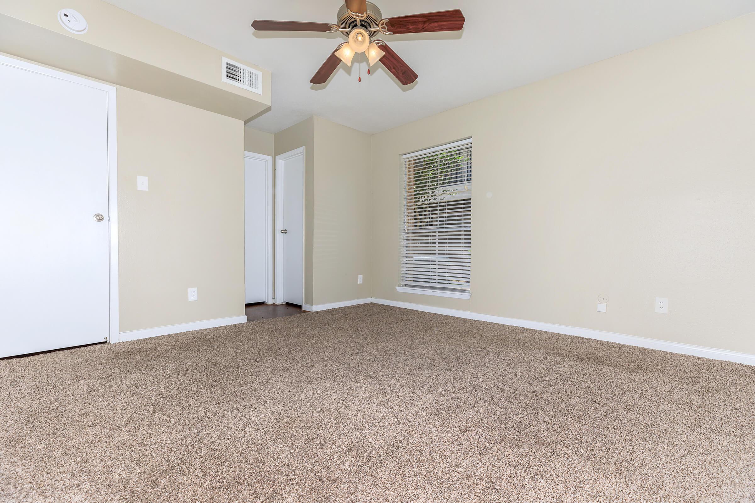 A well-lit empty room featuring beige walls, a ceiling fan with wooden blades, and plush carpet. There's a closed white door on the left and a second door in the background. A window with white blinds allows natural light to enter. The space appears clean and ready for furniture placement.