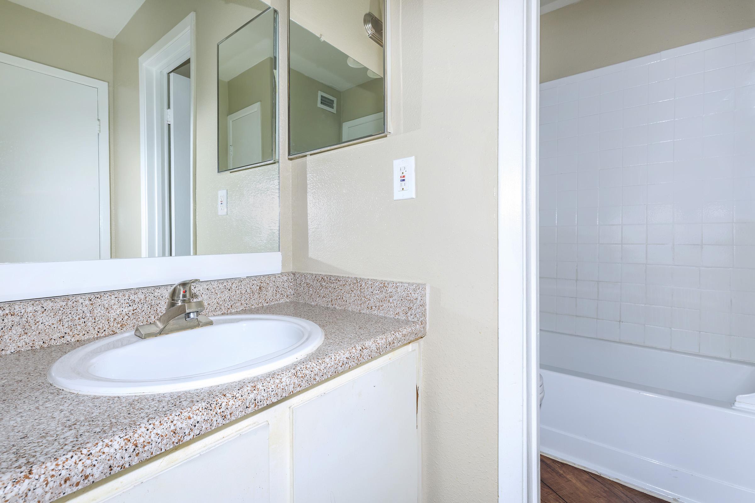 A clean, well-lit bathroom featuring a white sink with a granite countertop, a mirror above, and a bathtub with white tiles. The walls are painted in a soft beige color, and there is a doorway visible leading to another room. Natural light fills the space, enhancing its fresh appearance.