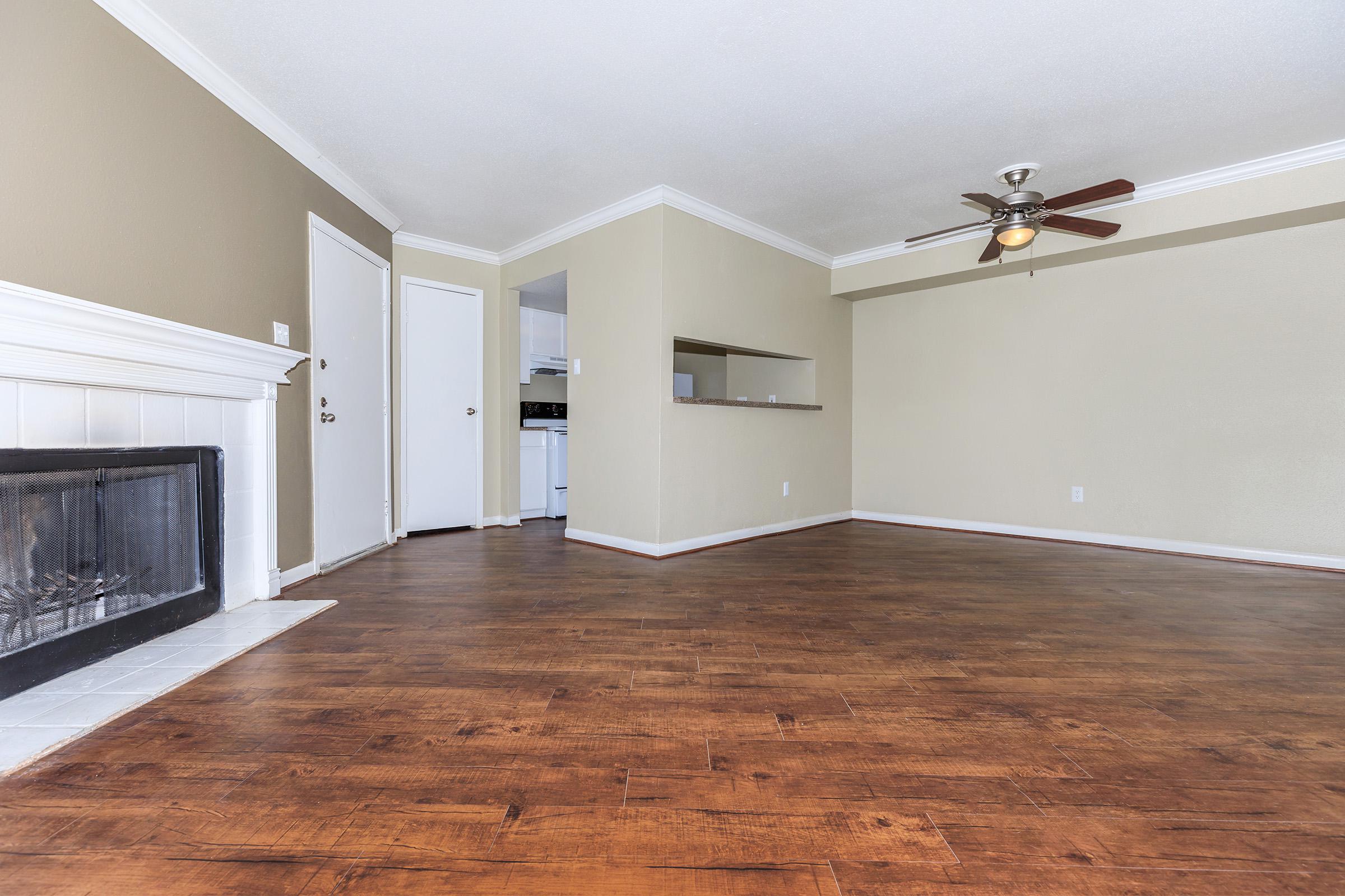 Empty living room with wooden flooring, a fireplace, and a ceiling fan. Walls are painted light beige, and there is a mirrored wall section. A doorway leads to a kitchen area in the background. The space is well-lit with natural light.