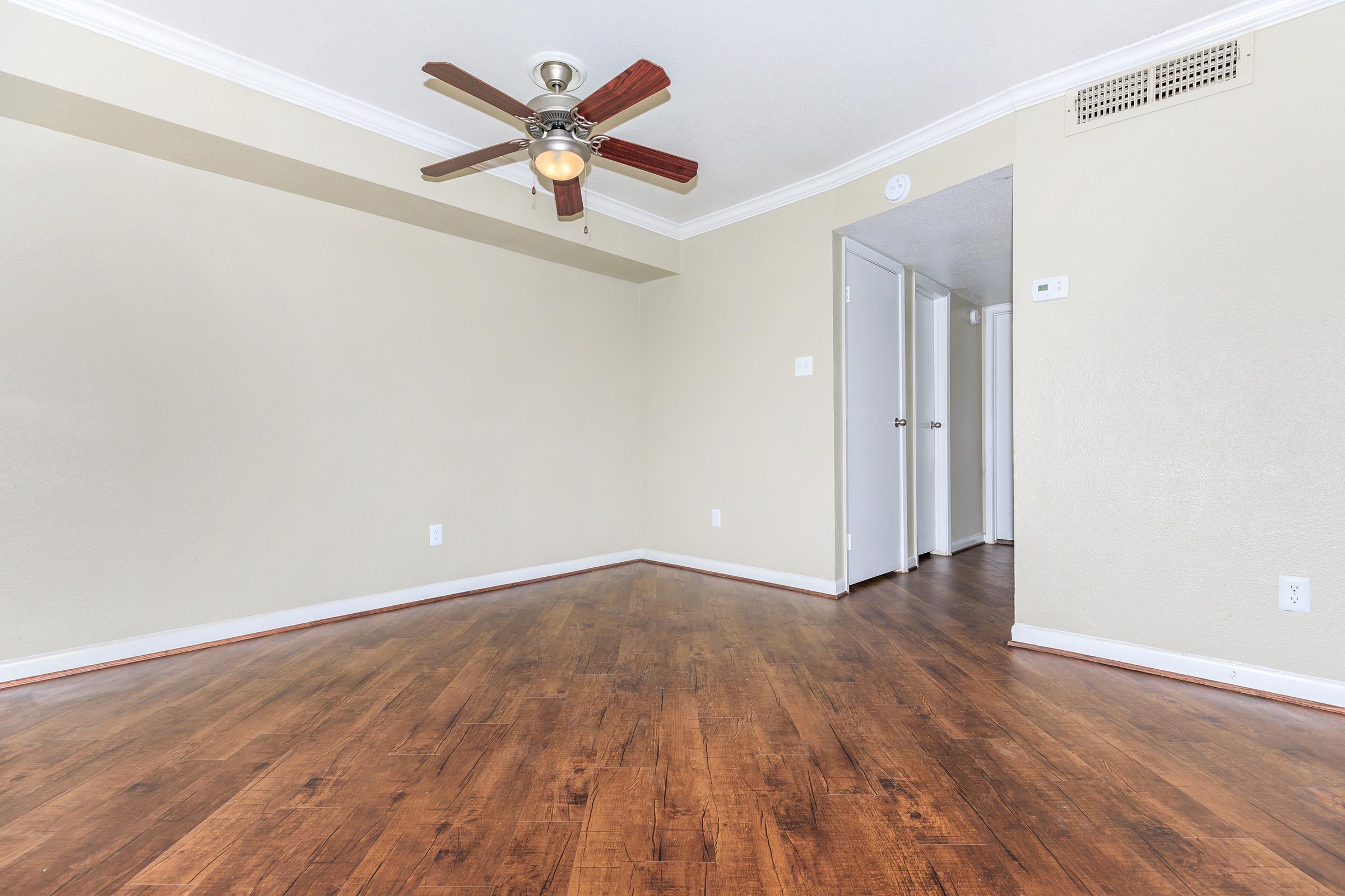 A spacious, unfurnished room with light-colored walls and hardwood flooring. A ceiling fan is mounted in the center, and there is a doorway leading to another room or hallway on the right. The space is well-lit, providing a neutral backdrop for various decor options.