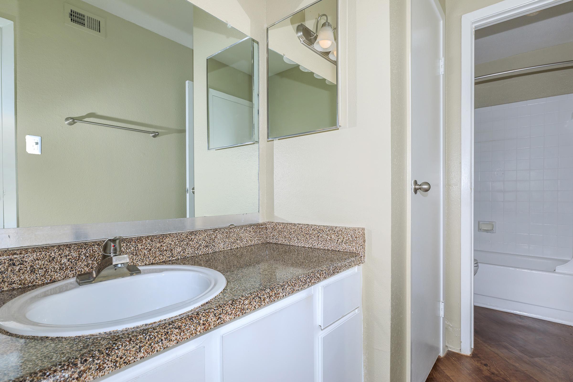 A clean bathroom featuring a white sink with a granite countertop, two mirrored cabinets above, and a light fixture. The walls are painted a light color, and there is a door leading to a shower area visible in the background. The floor has hardwood-like flooring.