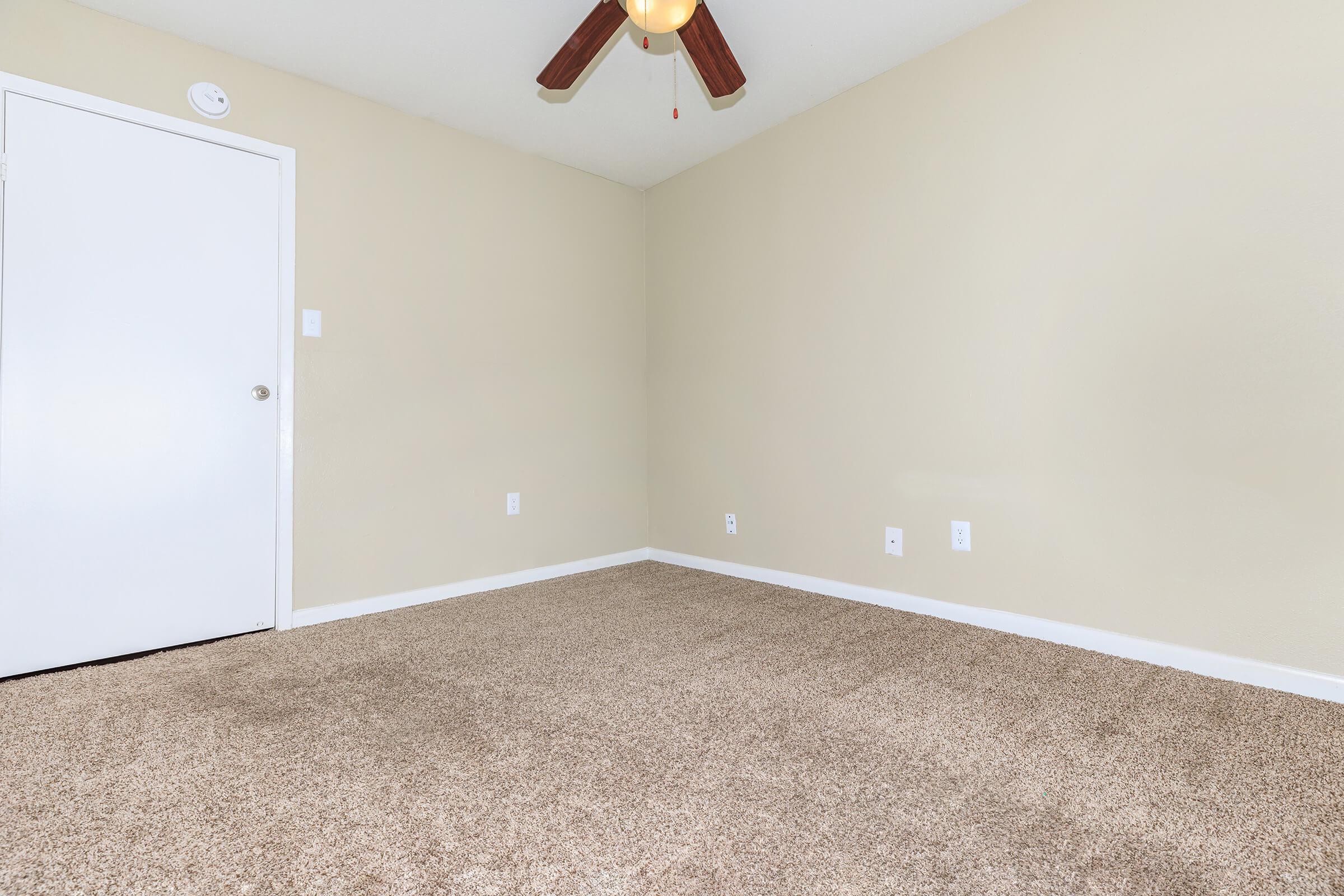 A blank, empty room featuring light beige walls and a ceiling fan with wooden blades. The floor is covered with light brown carpet. A white door is visible on the left side, and there are two small electrical outlets on the wall. The space appears bright and unoccupied.