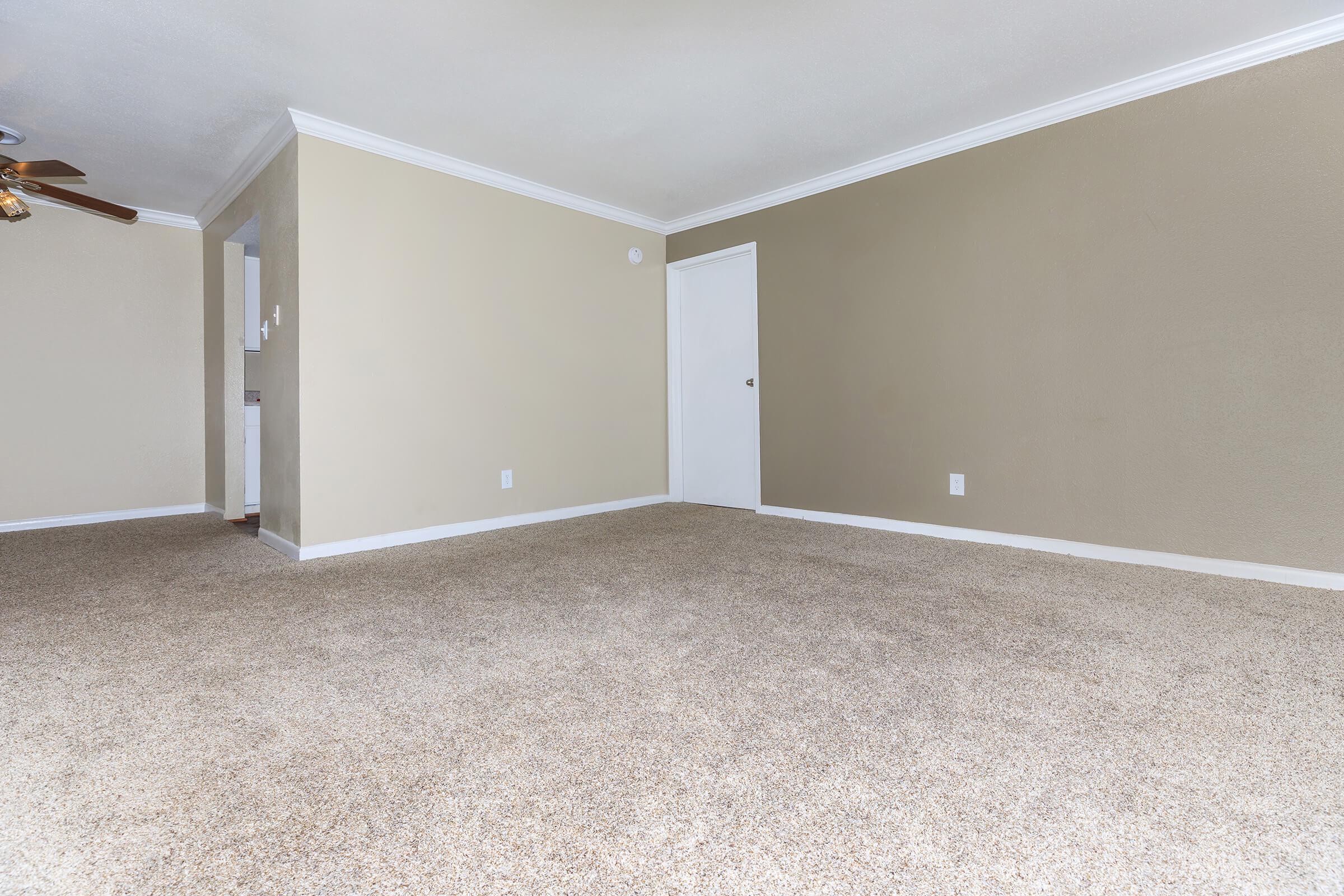 A spacious, empty living room featuring beige walls and light-colored carpet flooring. There is a ceiling fan visible on the left, and a closed white door on the right. Natural light illuminates the room, emphasizing its openness.