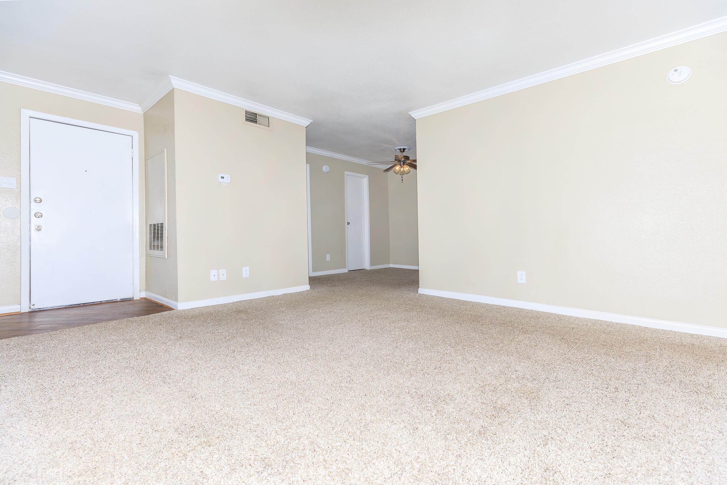 A spacious, unoccupied room with beige walls and light brown carpet, featuring a subtle ceiling fan. A white door leads to the outside, and another door is visible on the opposite wall. The room is well-lit and has a clean, minimalist appearance, ideal for various layouts and decor styles.