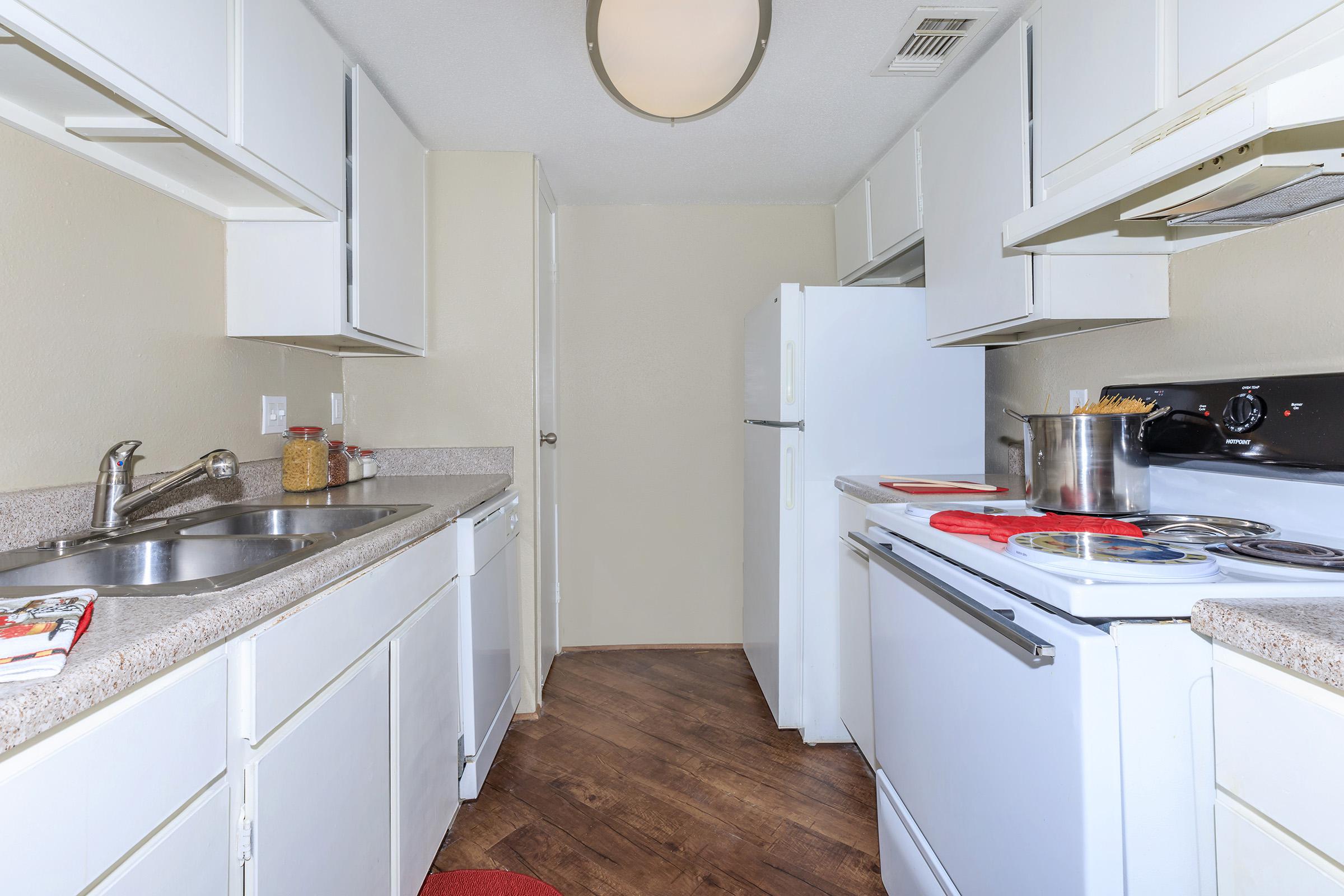 A well-lit kitchen featuring white cabinets and appliances, a stainless steel sink, and a pot on the stove. The countertops are beige, and there is a red kitchen mat on the floor. The space has wooden flooring and a doorway leading to another area.