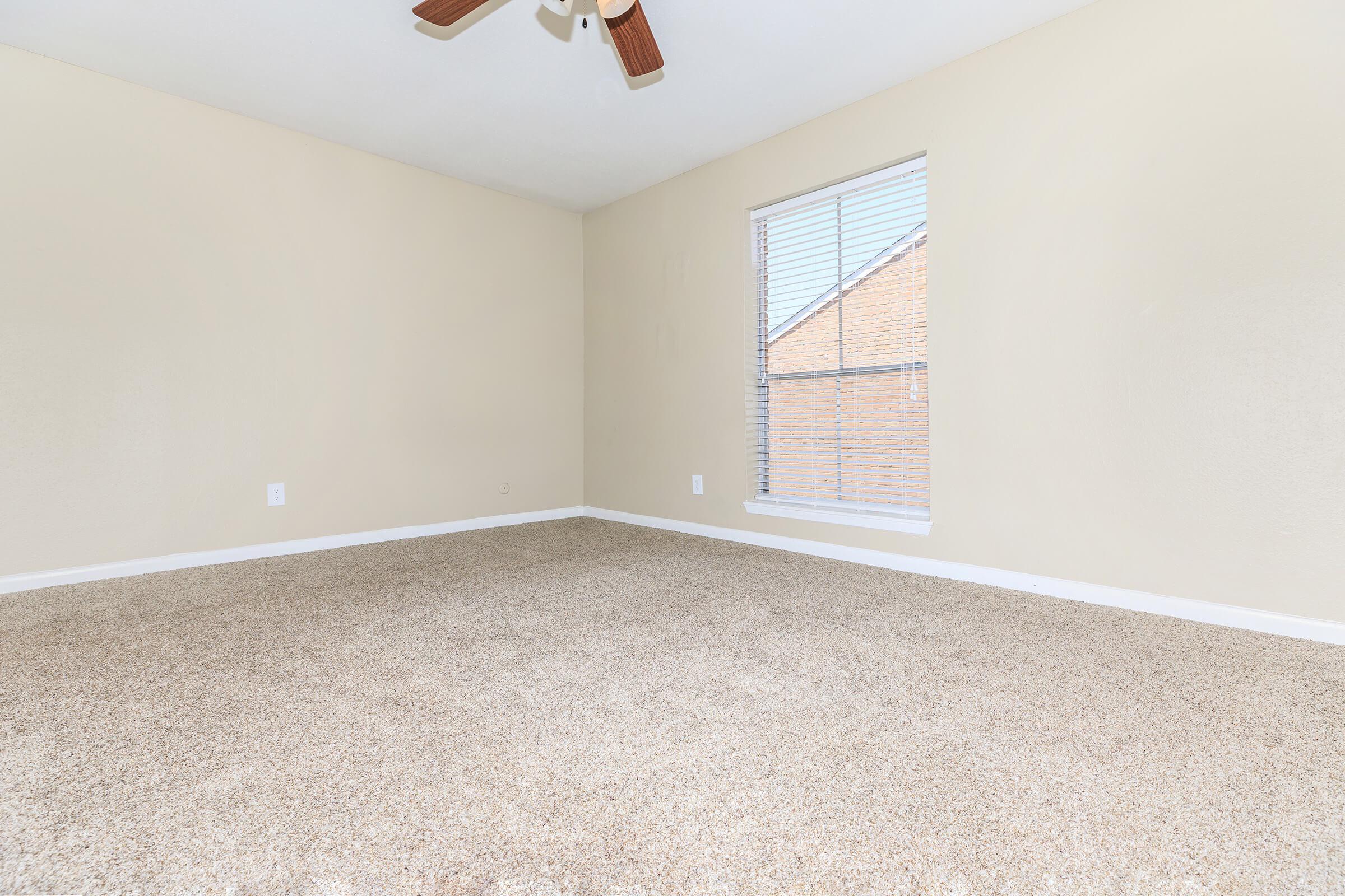 A spacious, empty room with light beige walls and a ceiling fan. The floor is covered in soft, tan carpet. A window with blinds allows natural light to enter, illuminating the space. The overall atmosphere is clean and bright, suitable for various uses.