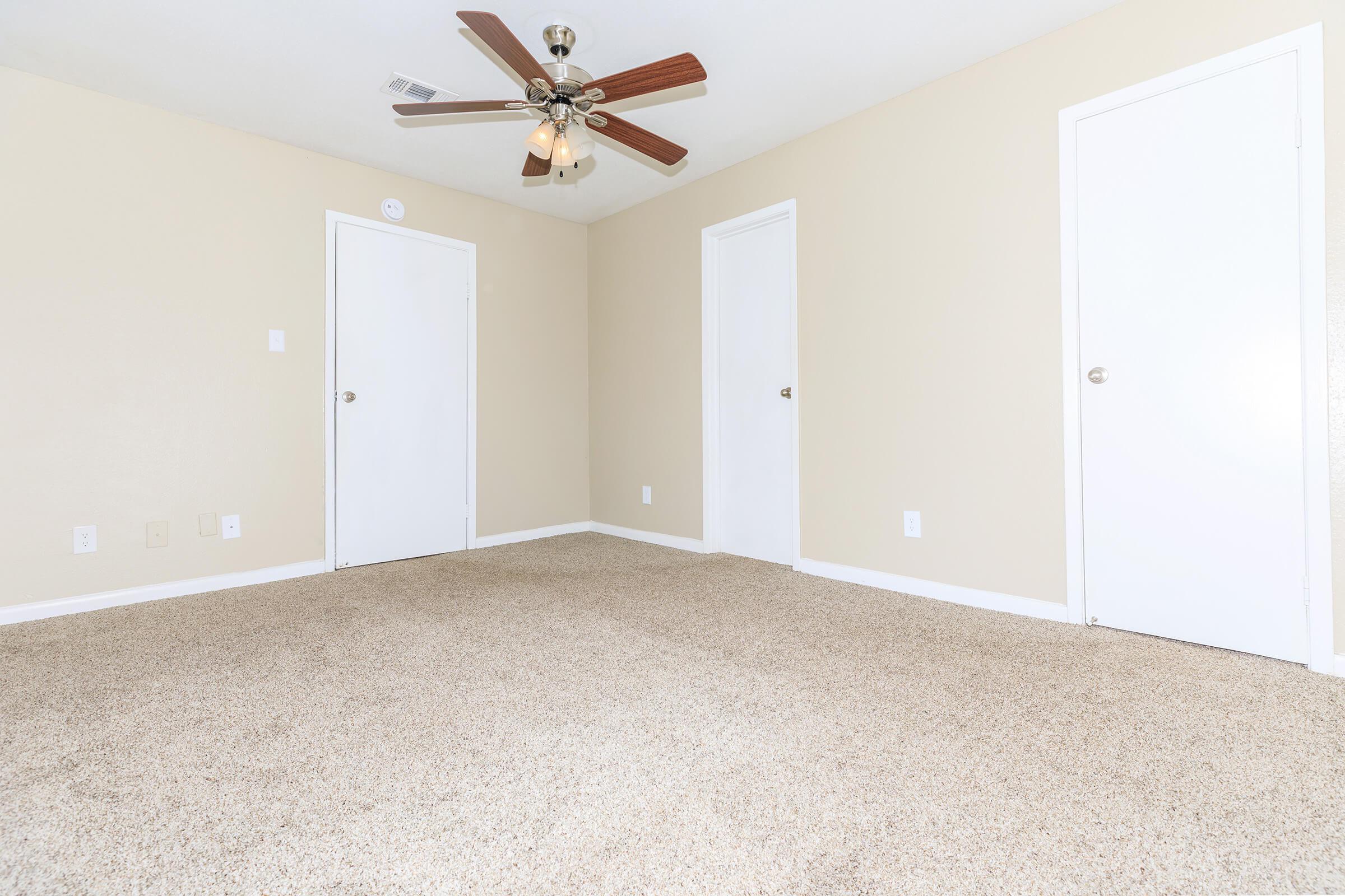 A well-lit, empty room featuring light beige walls and a ceiling fan with wooden blades. The floor is covered in beige carpet, and there are two white doors against the walls, providing a spacious and clean appearance.