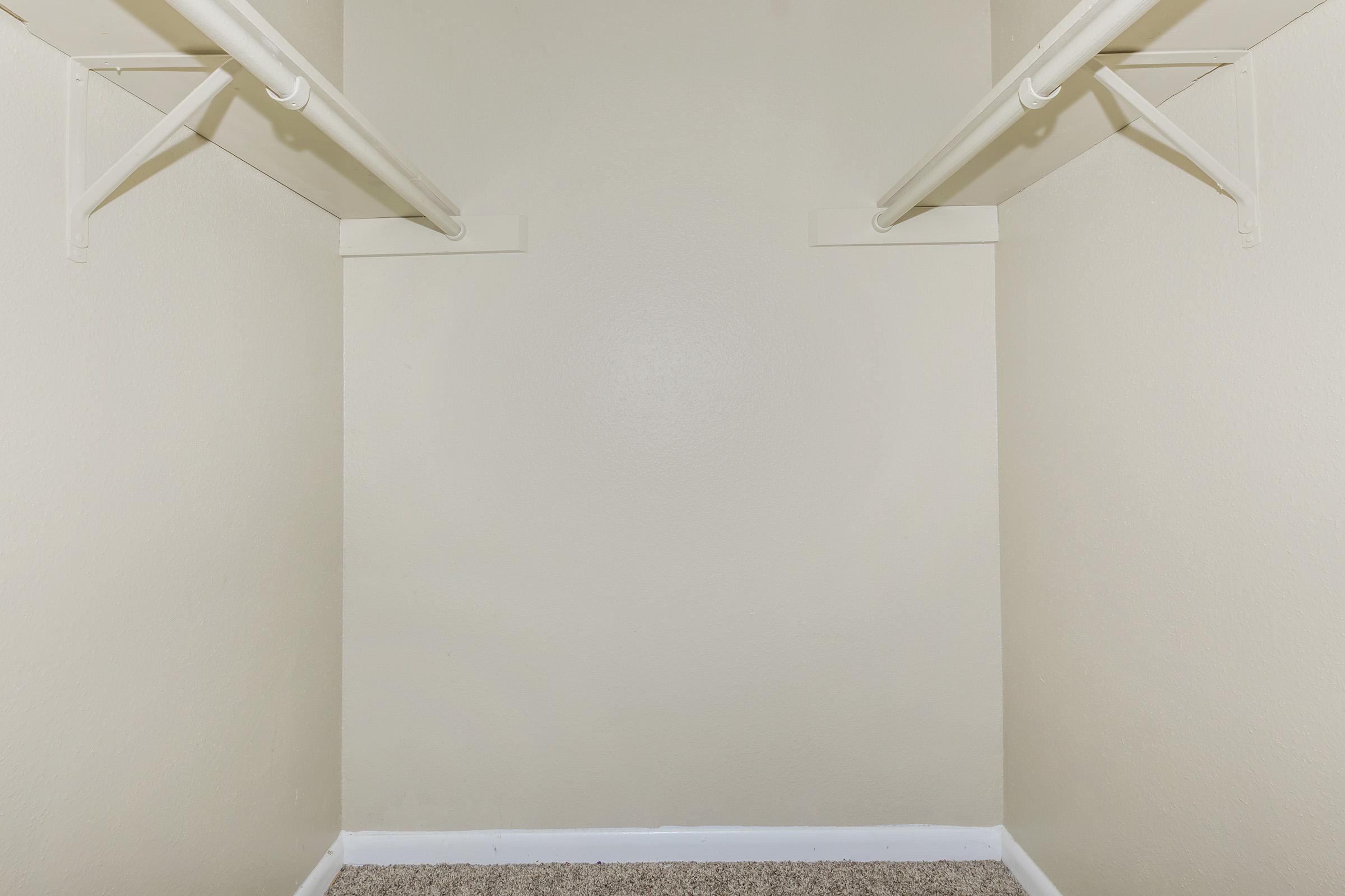 A vacant closet with two white hanging rods on opposite walls, and light beige walls. The floor is covered in a soft gray carpet, creating a simple and uncluttered appearance.