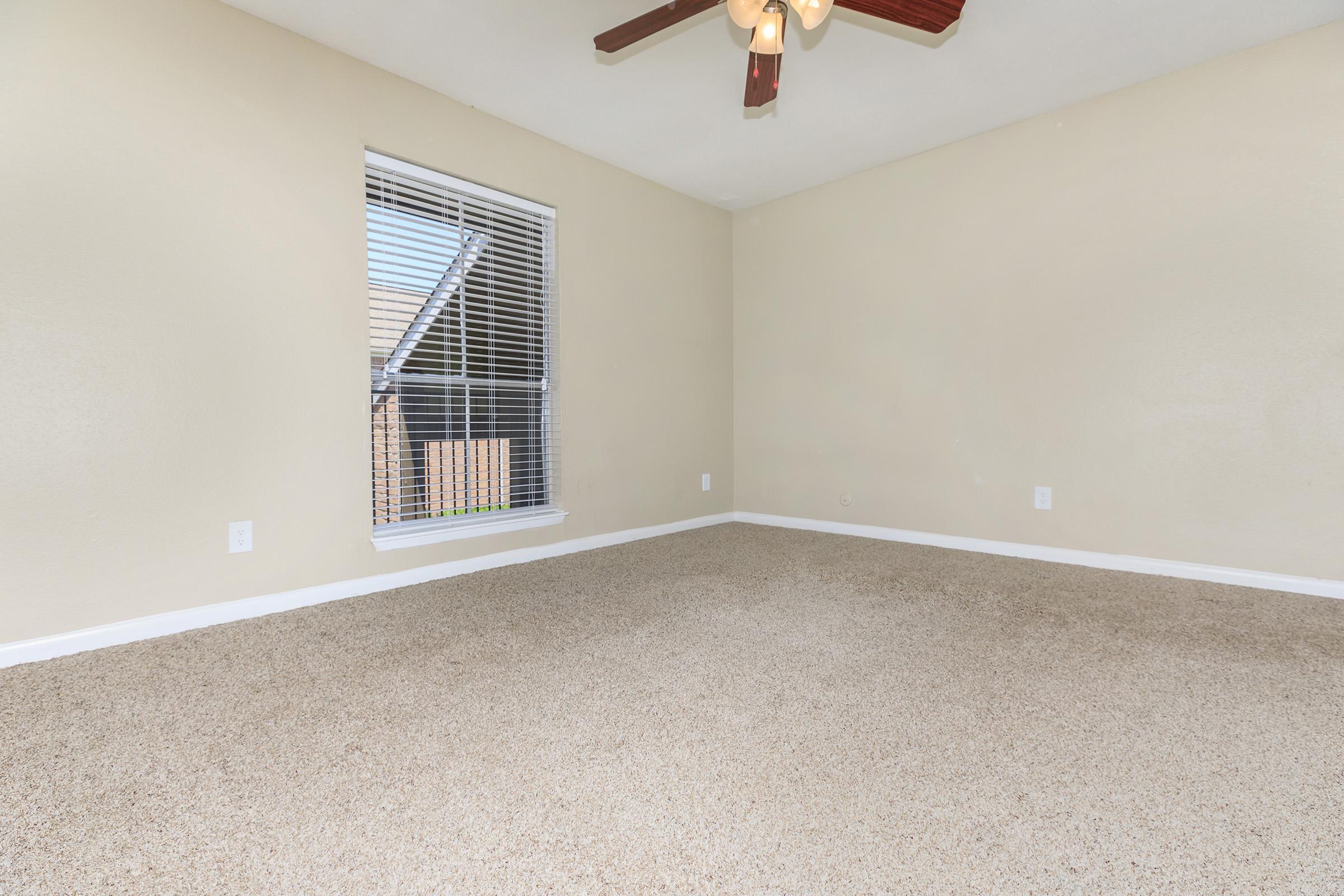 A spacious, empty room with light beige walls and a ceiling fan. The floor is covered in light brown carpet. A window with white blinds lets in natural light, providing a view of an outdoor area. The room has no furniture, emphasizing its openness and potential for various uses.