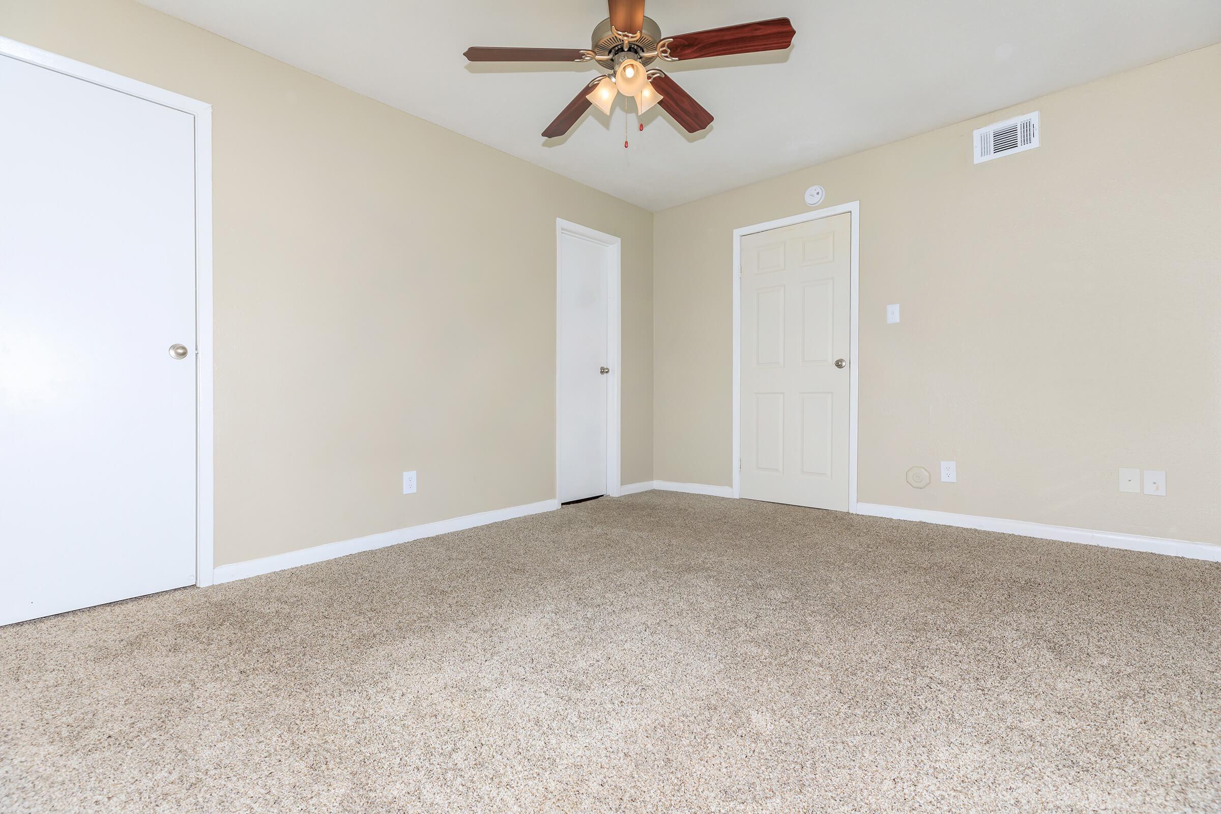 A spacious, light-colored room with beige walls and a ceiling fan featuring wooden blades. The carpet is a soft, light tan color. There are two doors: one leading to another room and another to a closet. The overall atmosphere is clean and inviting, with minimal furniture or decorations visible.