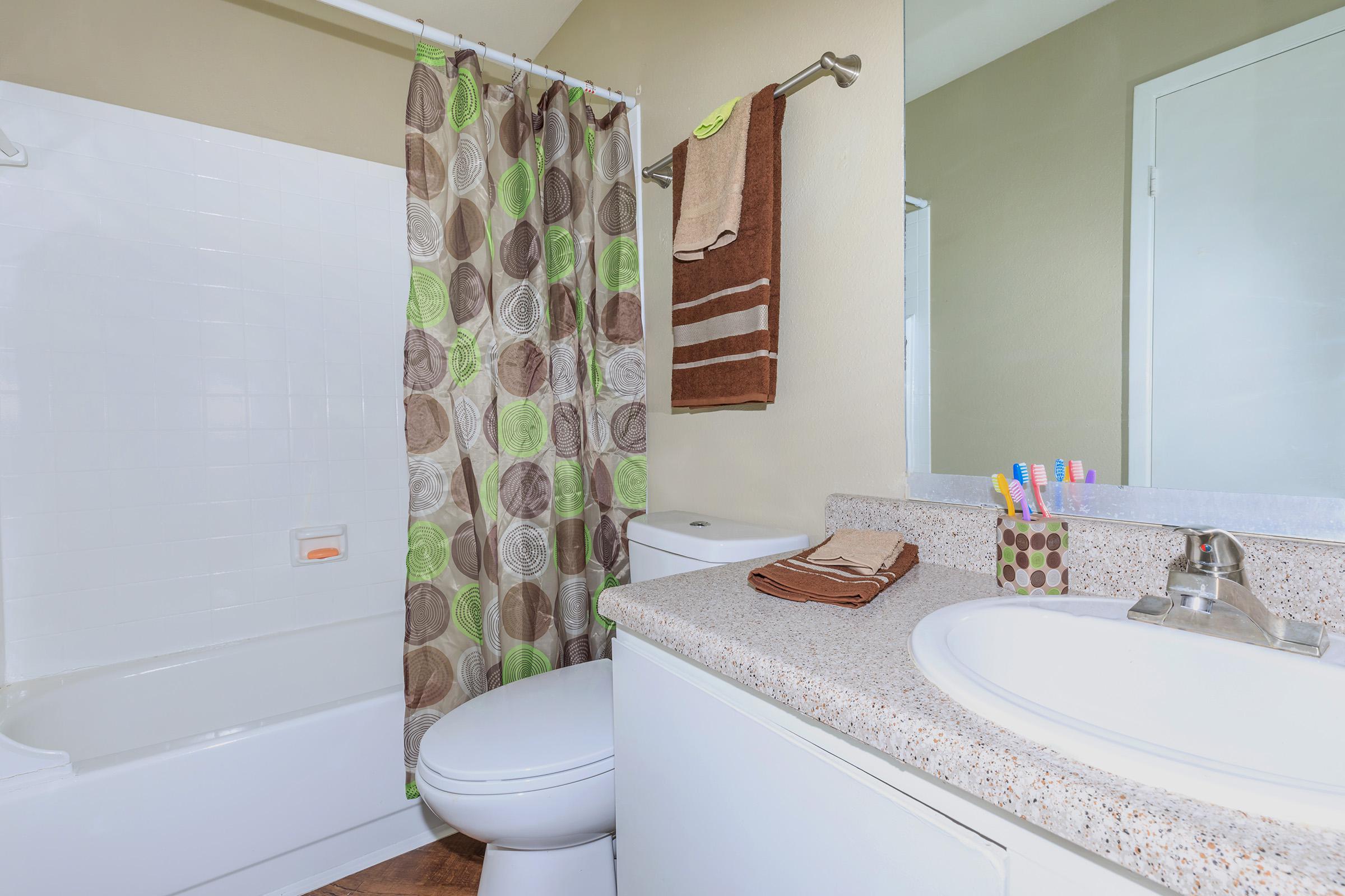 A clean and modern bathroom featuring a white bathtub with a shower curtain in green and brown circles. The countertop has a sink, towels, and toiletries, while there are decorative towels and a toothbrush holder nearby. The walls are painted in a neutral color, creating a bright and inviting atmosphere.