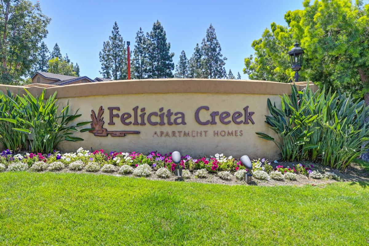 Sign for Felicita Creek Apartment Homes, featuring the name prominently displayed in a decorative font. The sign is set against a well-maintained landscape with colorful flowers and greenery, and there are trees in the background, indicating a pleasant residential environment.