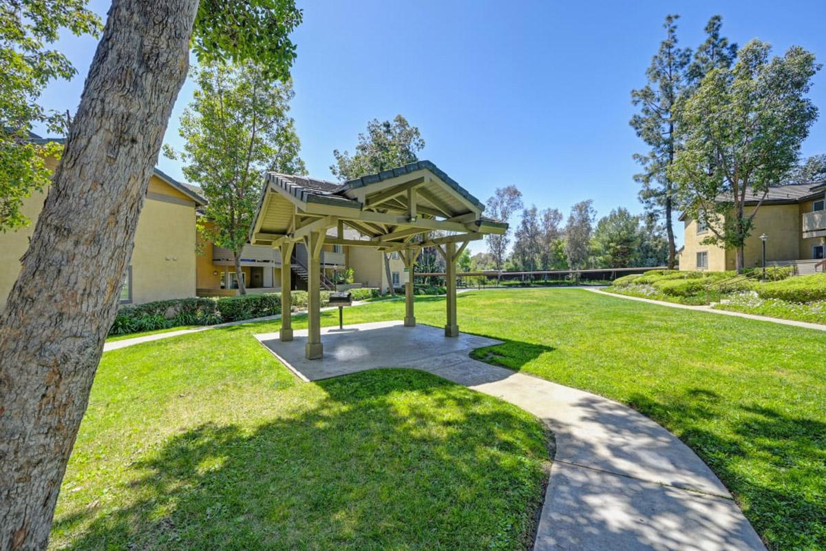 A grassy area featuring a wooden gazebo surrounded by trees and walkways, with two buildings visible in the background. The scene is bright and sunny, showcasing a well-maintained outdoor space for relaxation or gatherings.