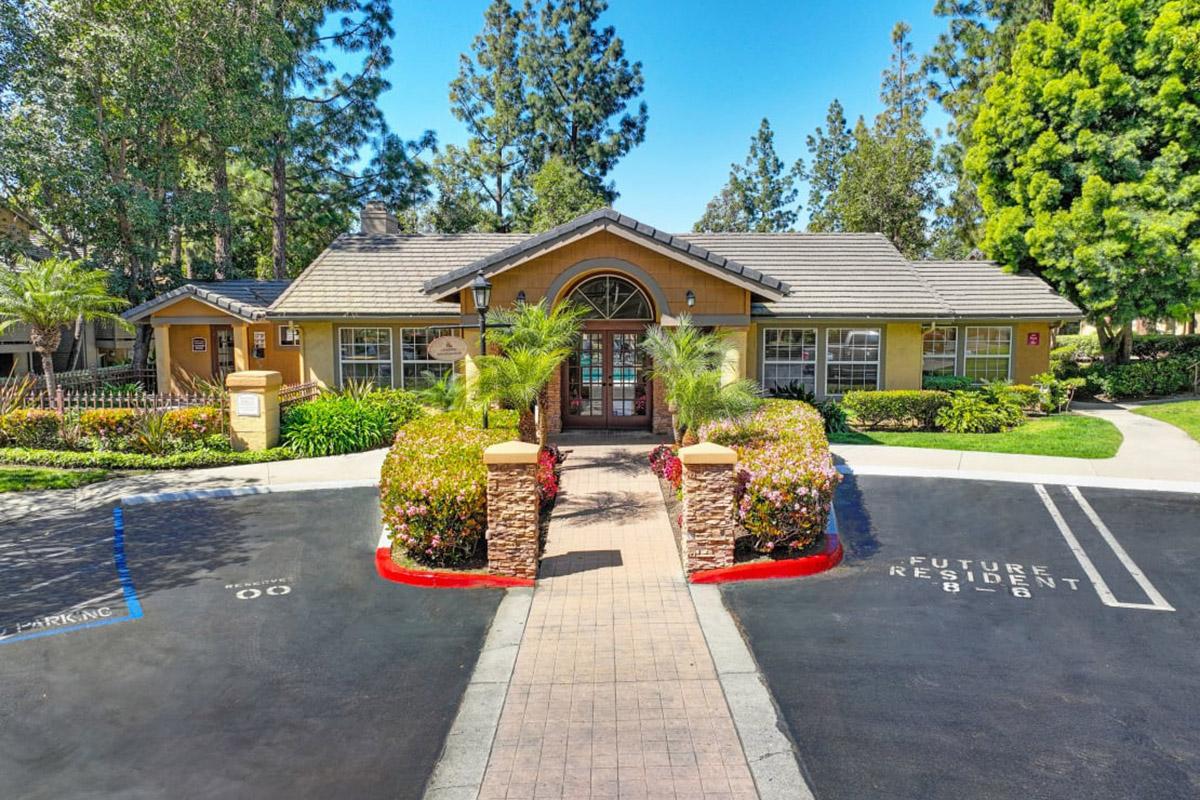 A welcoming residential building surrounded by lush greenery and colorful flowers. The pathway leads to the entrance, flanked by palm trees and well-maintained landscaping. Parking spaces are visible in front, with a designated area for future residents. The setting is bright and inviting, under a clear blue sky.
