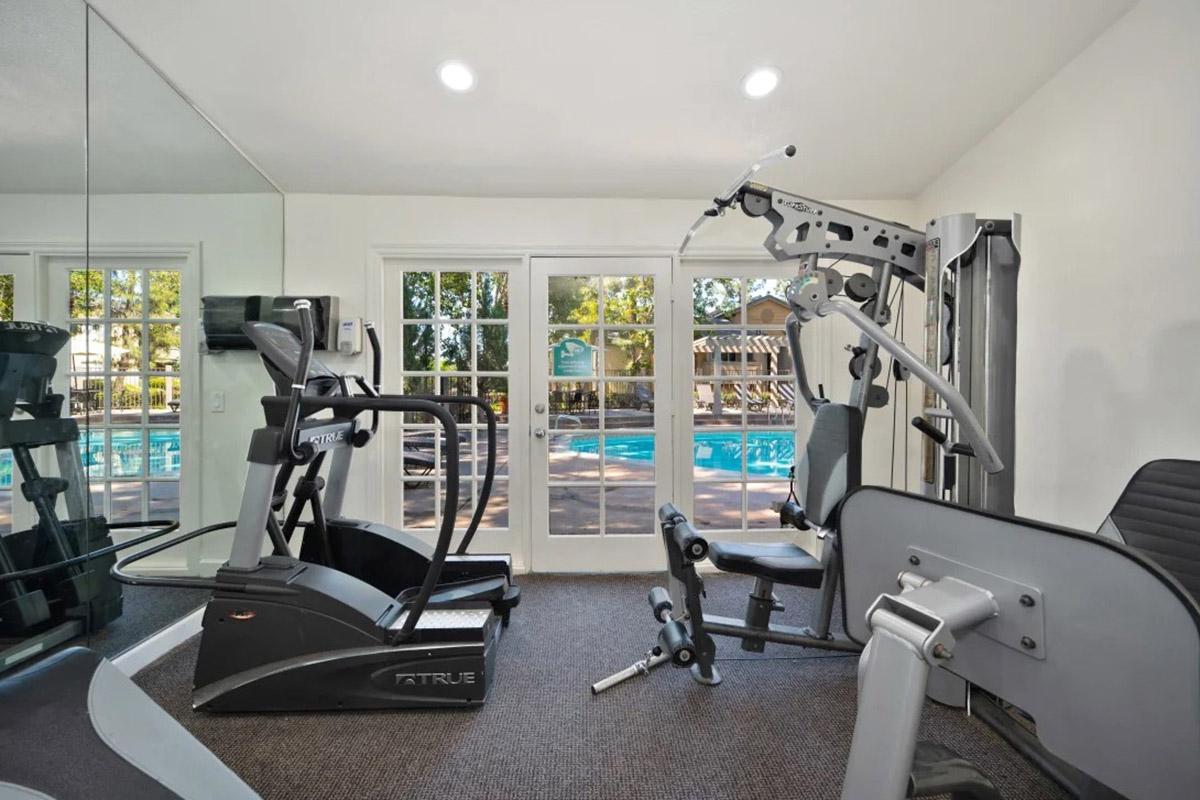 A well-lit gym interior featuring various fitness equipment, including an elliptical machine, a multi-station weight machine, and a workout bench. Large windows provide a view of an outdoor pool area, enhancing the inviting atmosphere.