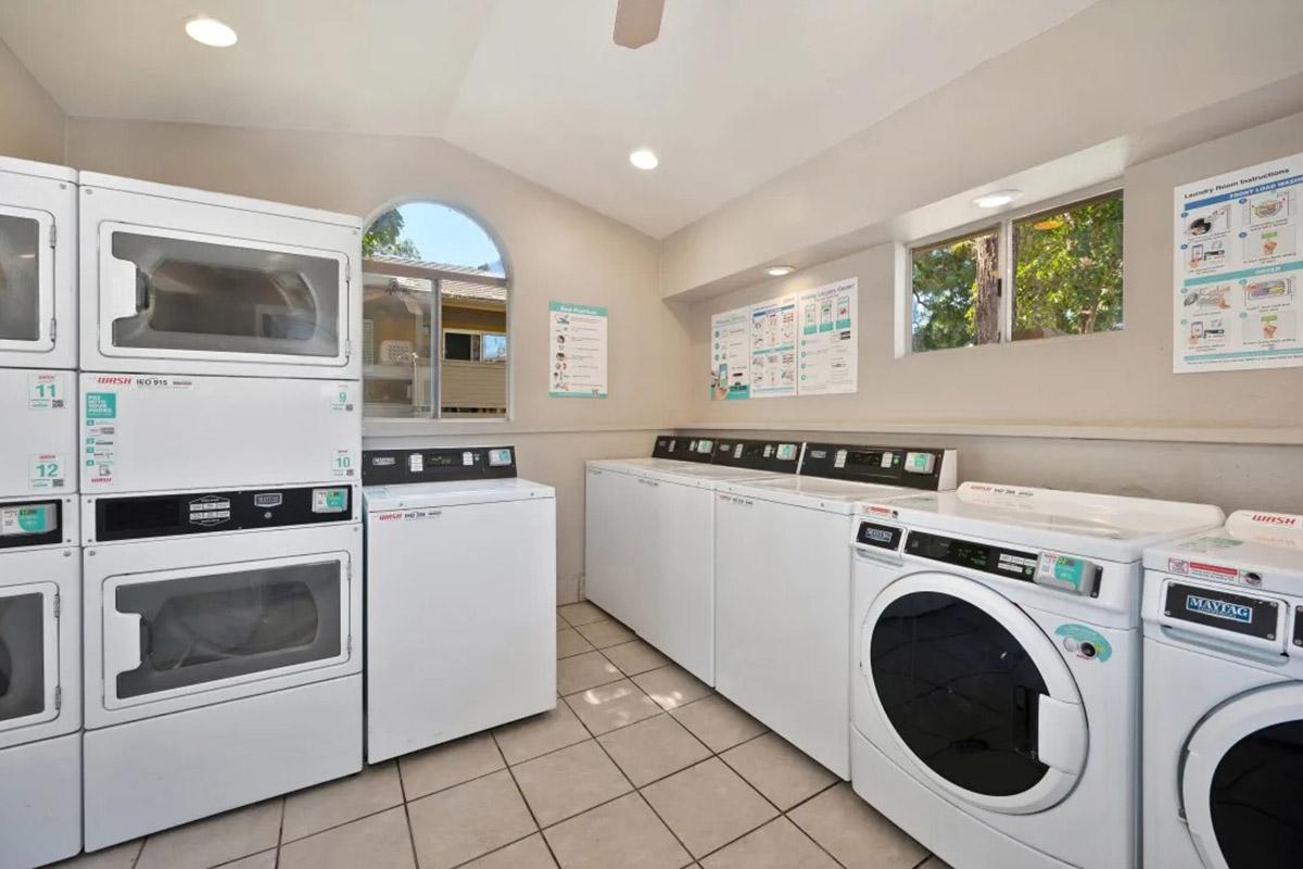 A clean and well-lit laundry room featuring several white washing machines and dryers arranged neatly along the walls, with informational posters on the sides. Large windows allow natural light to enter, enhancing the spacious atmosphere. The floor is tiled, and the overall decor is simple and functional.