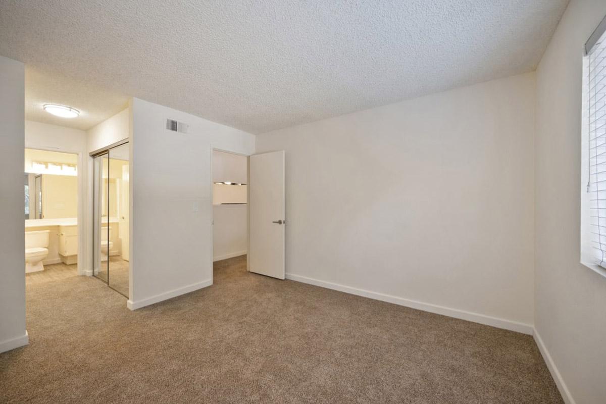 A spacious room featuring beige carpet, white walls, and a single door leading to a bathroom area. Natural light enters through a window, and there is a mirrored closet on one side. The layout is open, creating a feeling of ample space, with neutral colors and simple design.