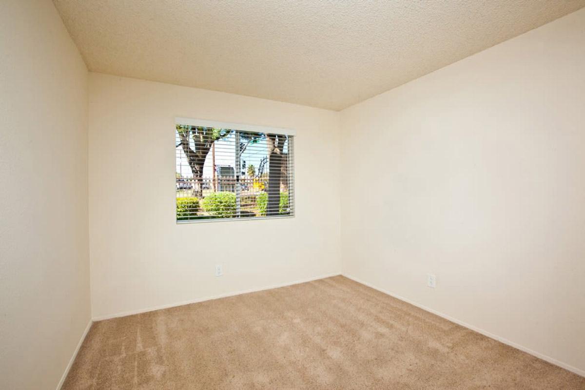 Empty room with beige walls and carpet. A window with blinds is visible, showing a view of greenery outside. The room is well-lit, with no furniture or decorations. The overall atmosphere is clean and spacious, suitable for use as a bedroom or office.