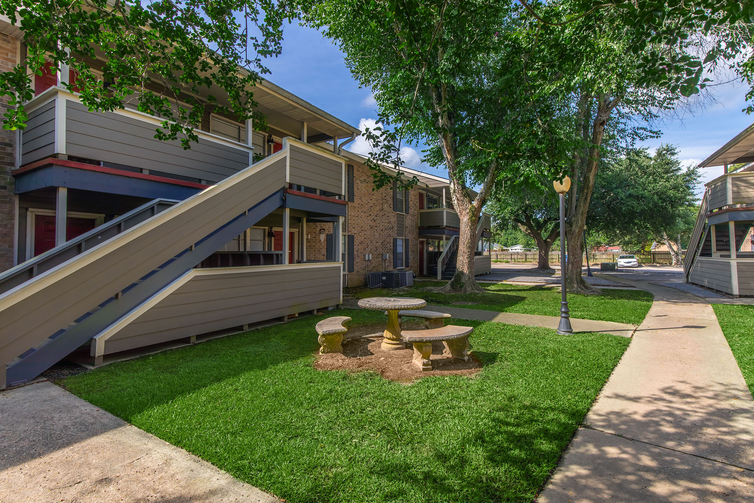 A well-maintained residential complex featuring two-story apartments with wooden steps, surrounded by green grass and trees. There is a circular stone table with matching benches in the foreground, and well-defined walkways leading through the area. Bright blue skies add to the inviting atmosphere.