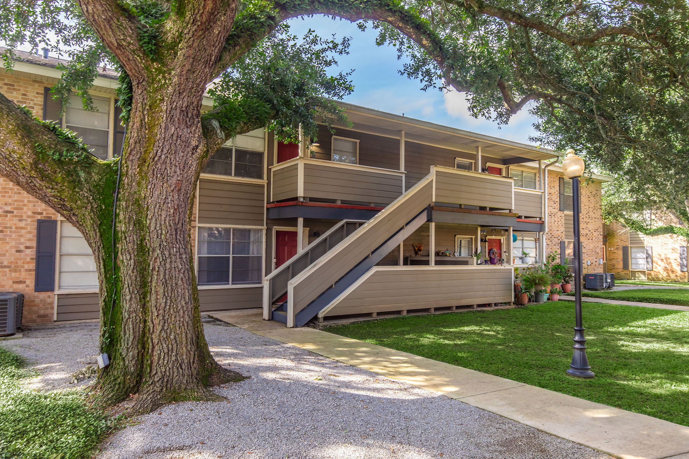 A well-maintained multi-unit apartment building with a wooden staircase and balcony. The building is surrounded by green grass, trees, and a pathway. There are decorative elements on the balcony and a lamppost nearby, creating an inviting atmosphere.