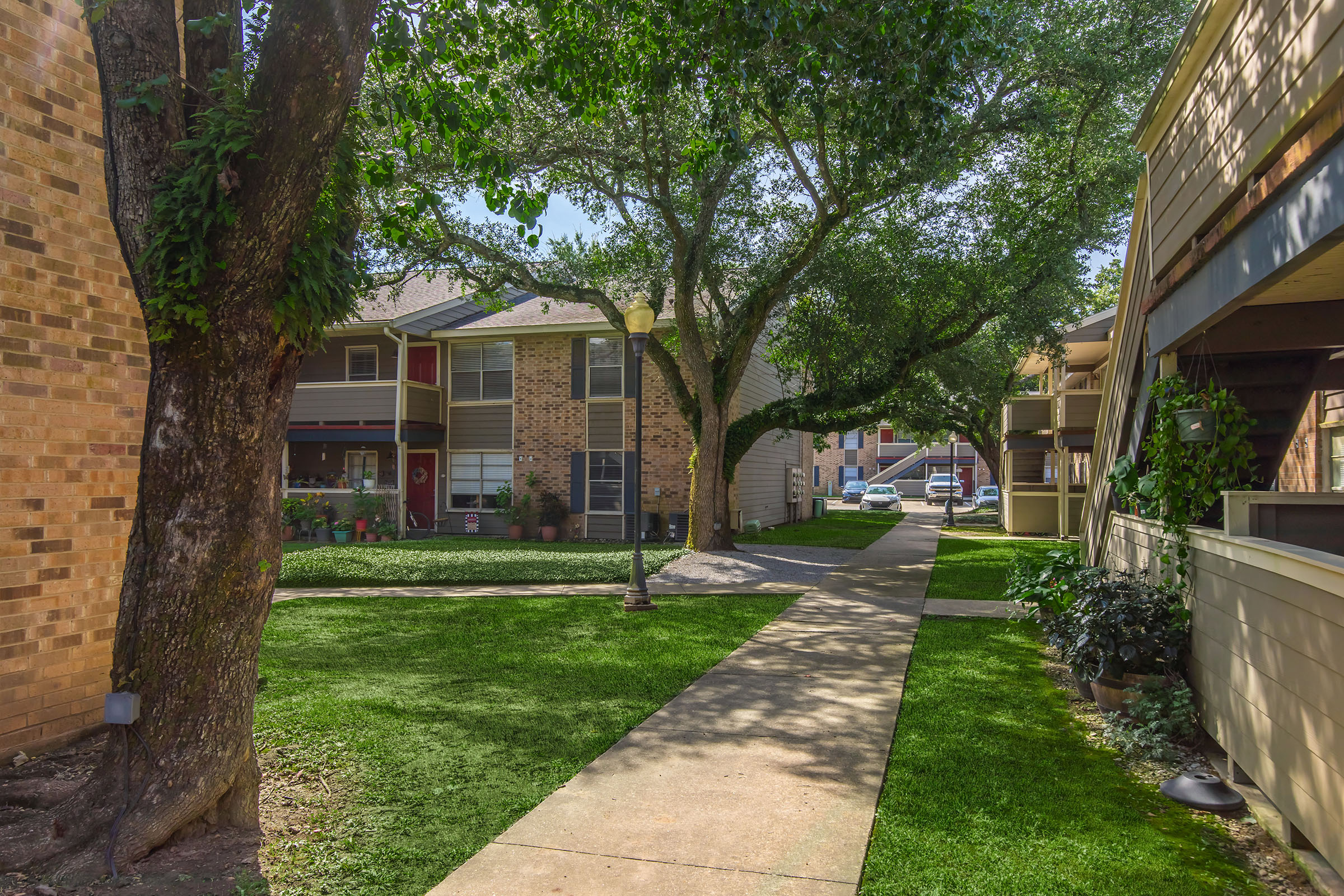 A view of an apartment complex with greenery, featuring two buildings on either side of a paved pathway. Lush trees and well-maintained lawns create a serene atmosphere. Cars are parked in the background, indicating residential activity. The scene is bright and inviting, suggesting a pleasant living environment.