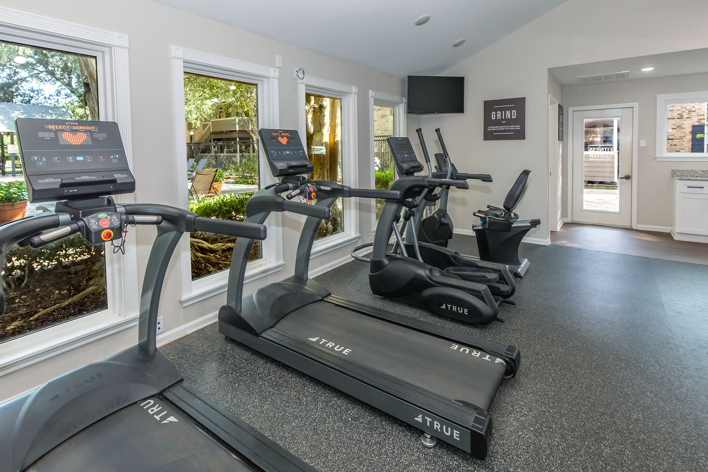 A modern gym space featuring several treadmills and exercise bikes, with large windows allowing natural light and a view of greenery outside. The room is equipped with fitness machines, a television, and motivational wall decor. The flooring is dark and rubberized for safety and comfort.
