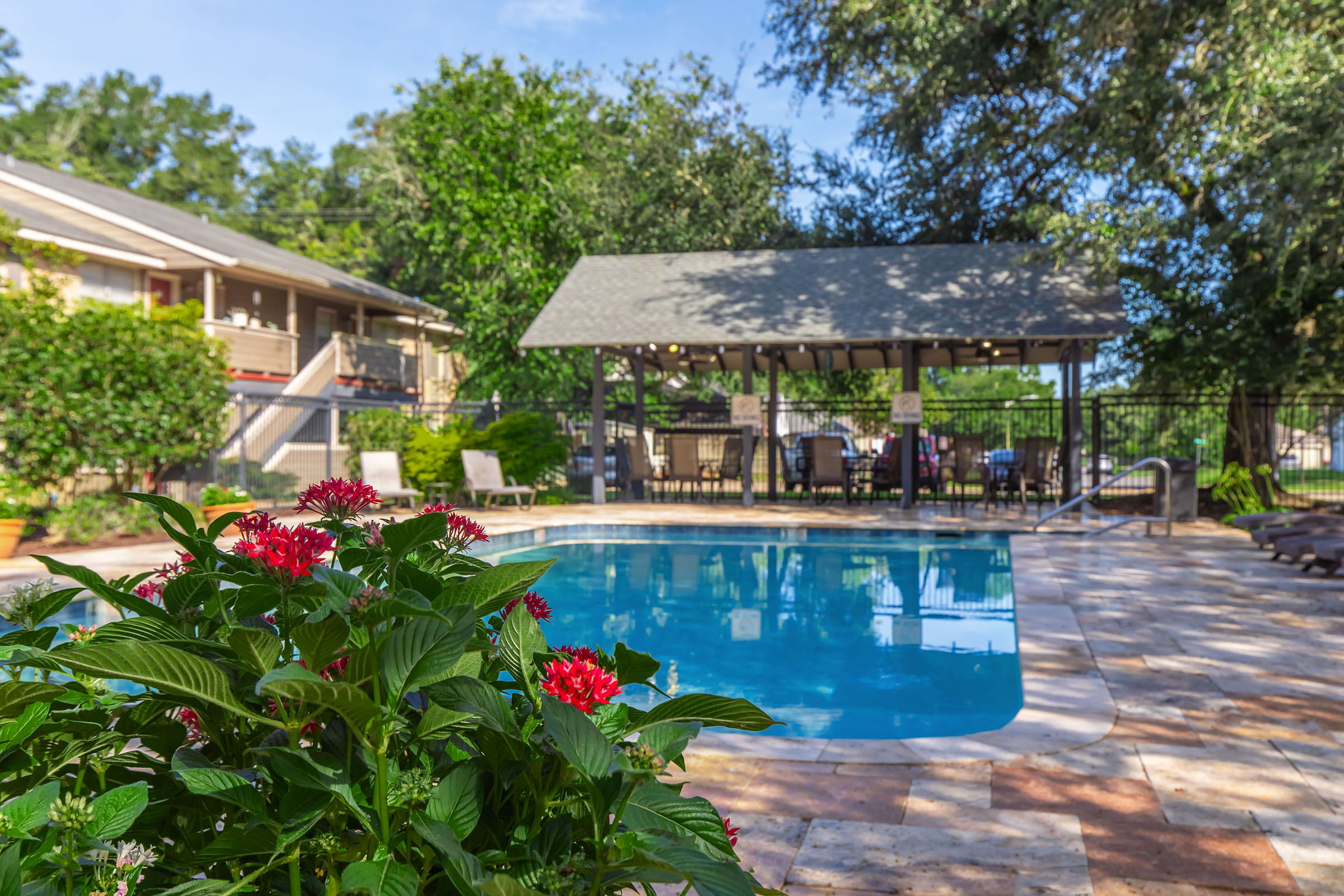 A tranquil outdoor pool area surrounded by lush greenery and vibrant flowers. The pool is clear and inviting, with a shaded seating area nearby under a gazebo. Comfortable lounge chairs are positioned around the pool, creating a relaxing atmosphere in a sunny environment.