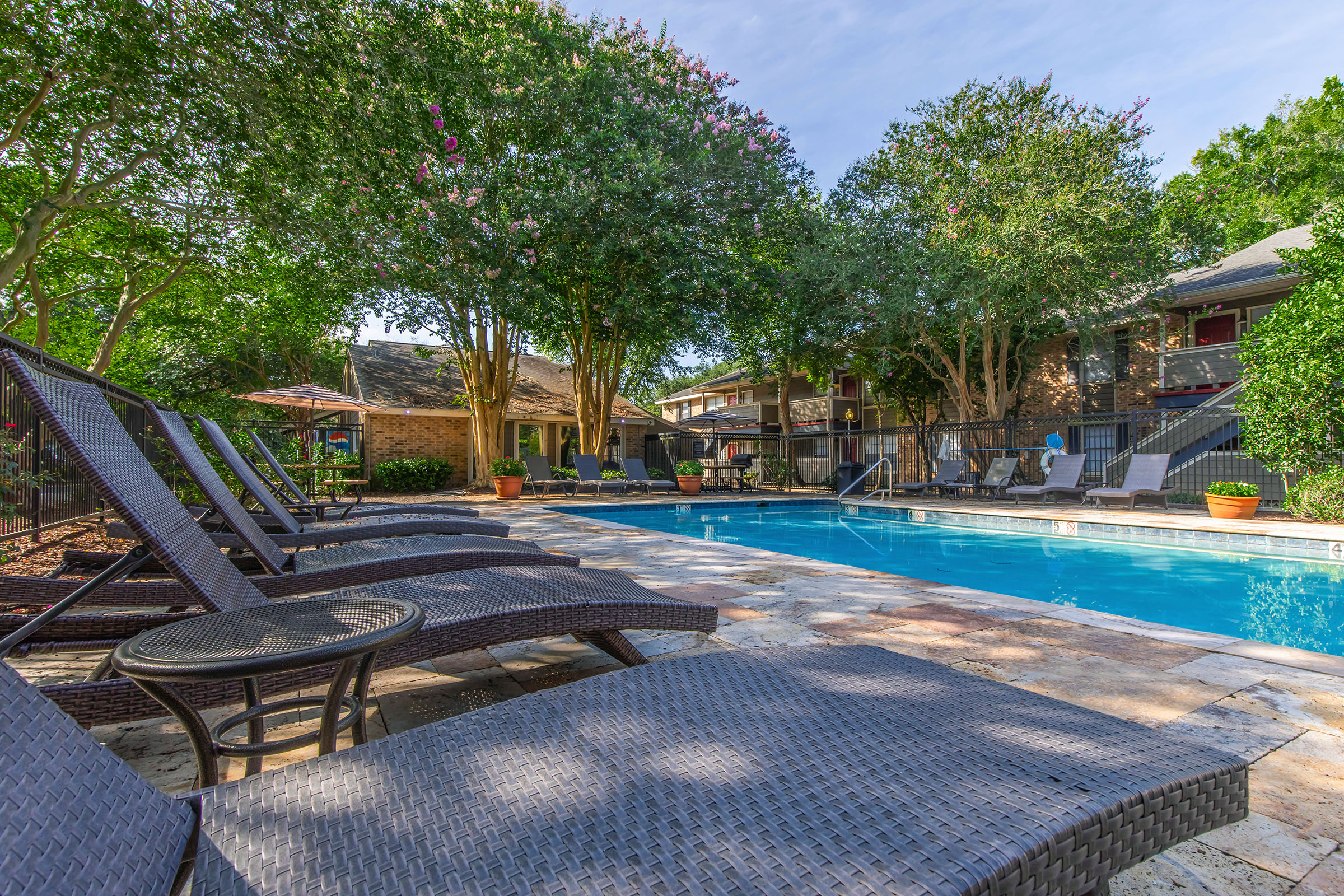 A tranquil outdoor scene featuring a swimming pool surrounded by lounge chairs and lush green trees. The pool area is designed for relaxation, with potted plants and a few shaded seating areas, creating a serene atmosphere perfect for leisure or summer gatherings.