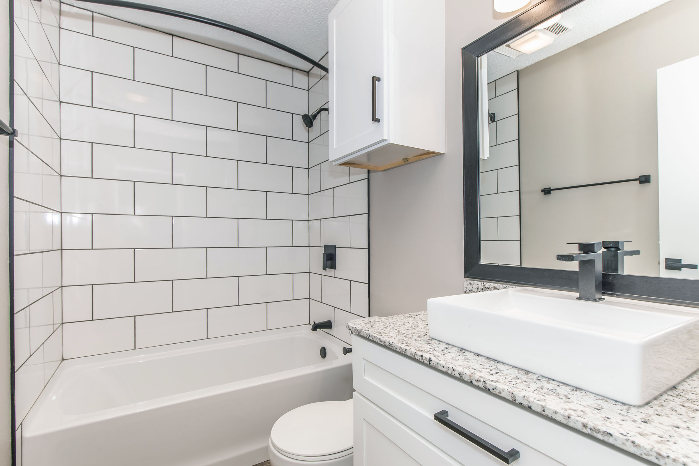 A modern bathroom featuring a white tiled shower and tub area, a countertop with a rectangular sink, and a large mirror above. The walls are painted gray, and there are black fixtures and accents throughout the space, providing a sleek and contemporary look.