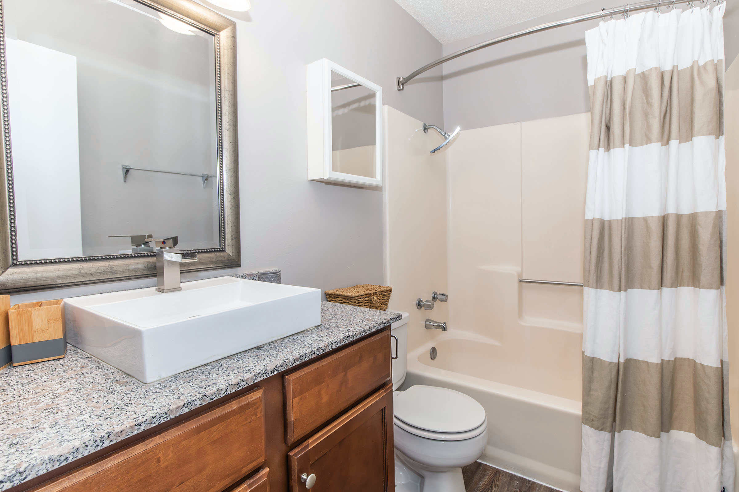 A clean and modern bathroom featuring a beige bathtub and shower combination, a stylish rectangular sink on a granite countertop, wooden cabinets, and a large mirror. The shower area has a striped curtain, and there is a small white cabinet mounted on the wall. A woven basket sits beside the sink.