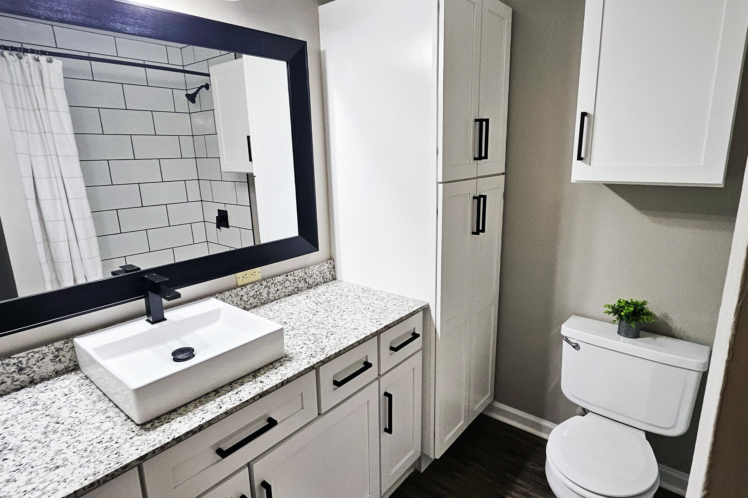 A modern bathroom featuring a white granite countertop with an integrated sink, a large mirror with a black frame, a tall white cabinet, and a toilet. The space has light gray walls and a shower area with a white curtain and tiled wall. A small plant adds a touch of greenery to the countertop.
