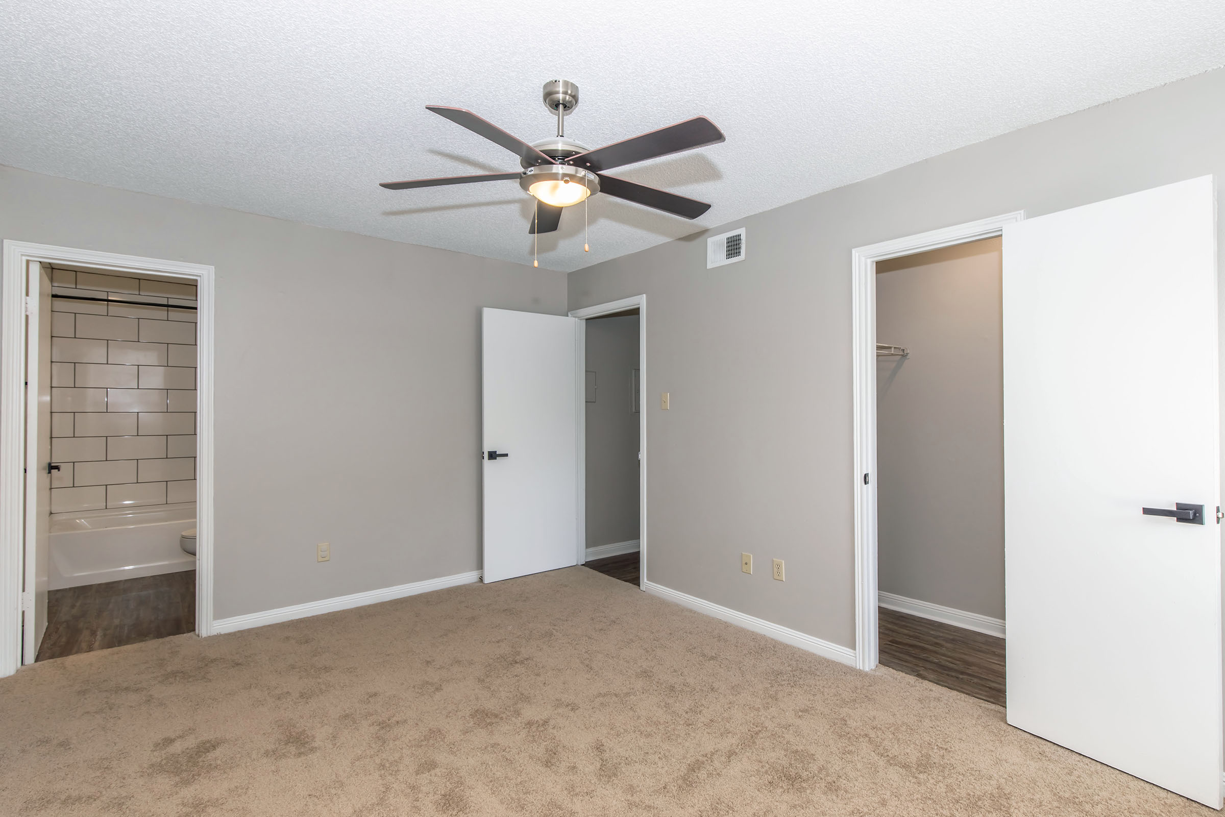 A neutral-colored bedroom with a ceiling fan and carpeted flooring. There are two open doors leading to other rooms, one of which appears to be a bathroom. The walls are painted light gray, and there's a small closet space visible near the bathroom door.