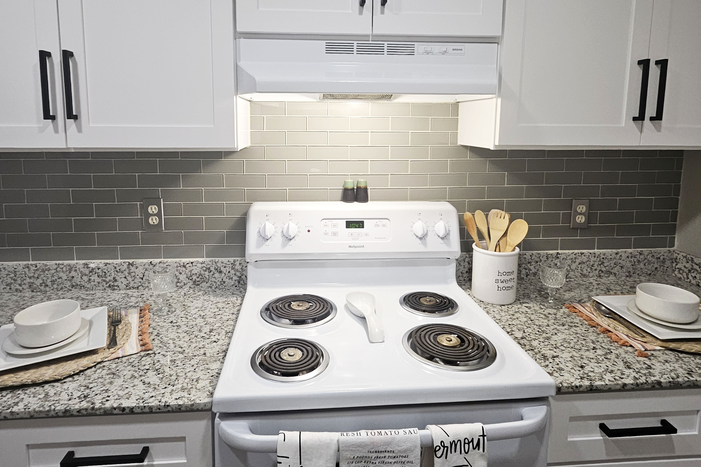 A clean kitchen countertop featuring a white stove with four burners, a light overhead, and a grey tiled backsplash. Two plates and bowls are set on woven placemats, with wooden utensils in a container nearby. The countertop is made of speckled grey and white granite, creating a modern and inviting cooking space.