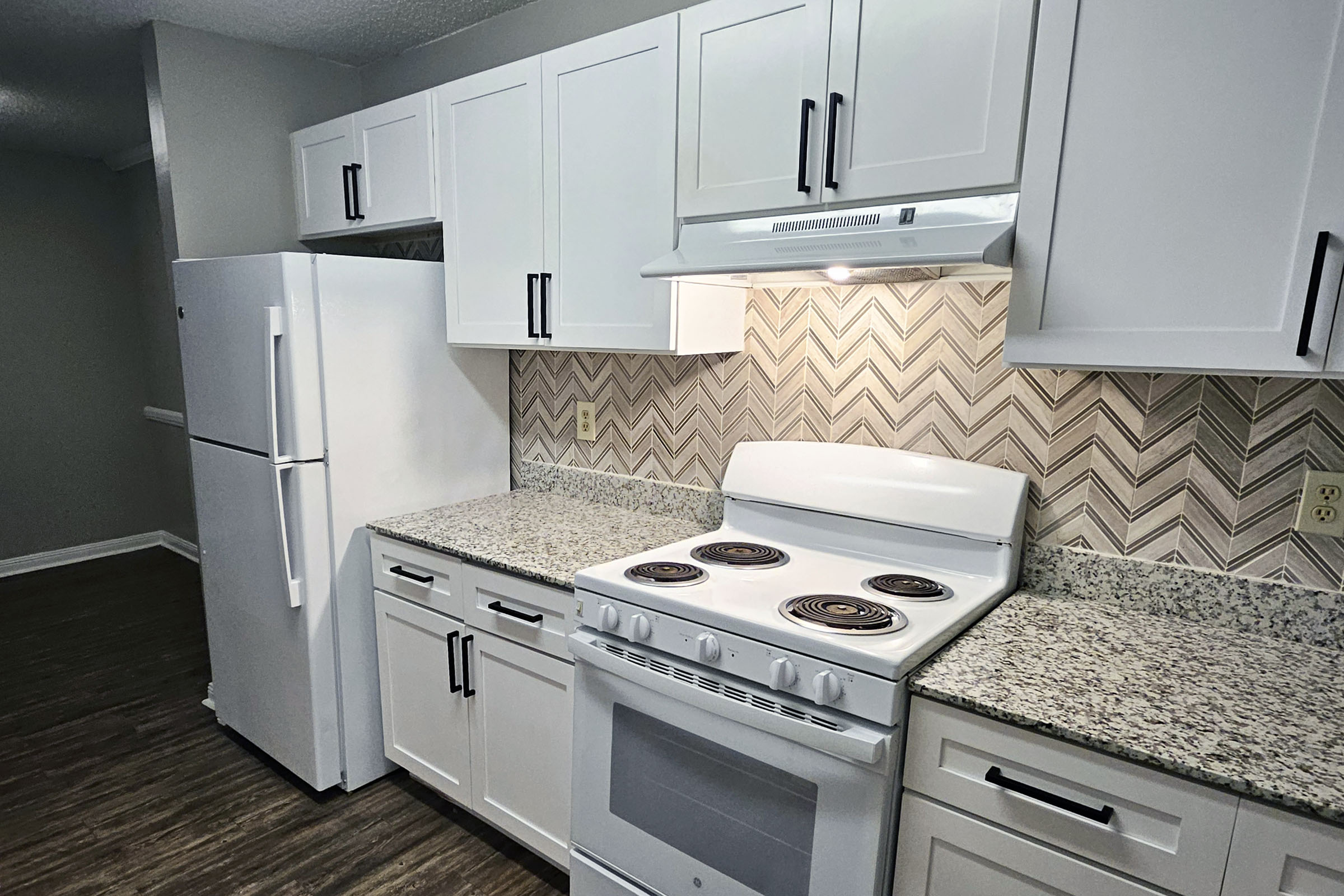 Modern kitchen featuring white cabinetry, a granite countertop, and a white gas stove with four burners. The space includes a white refrigerator, an overhead range hood, and beige chevron-patterned backsplash tiles, all set against a neutral wall and wooden flooring.