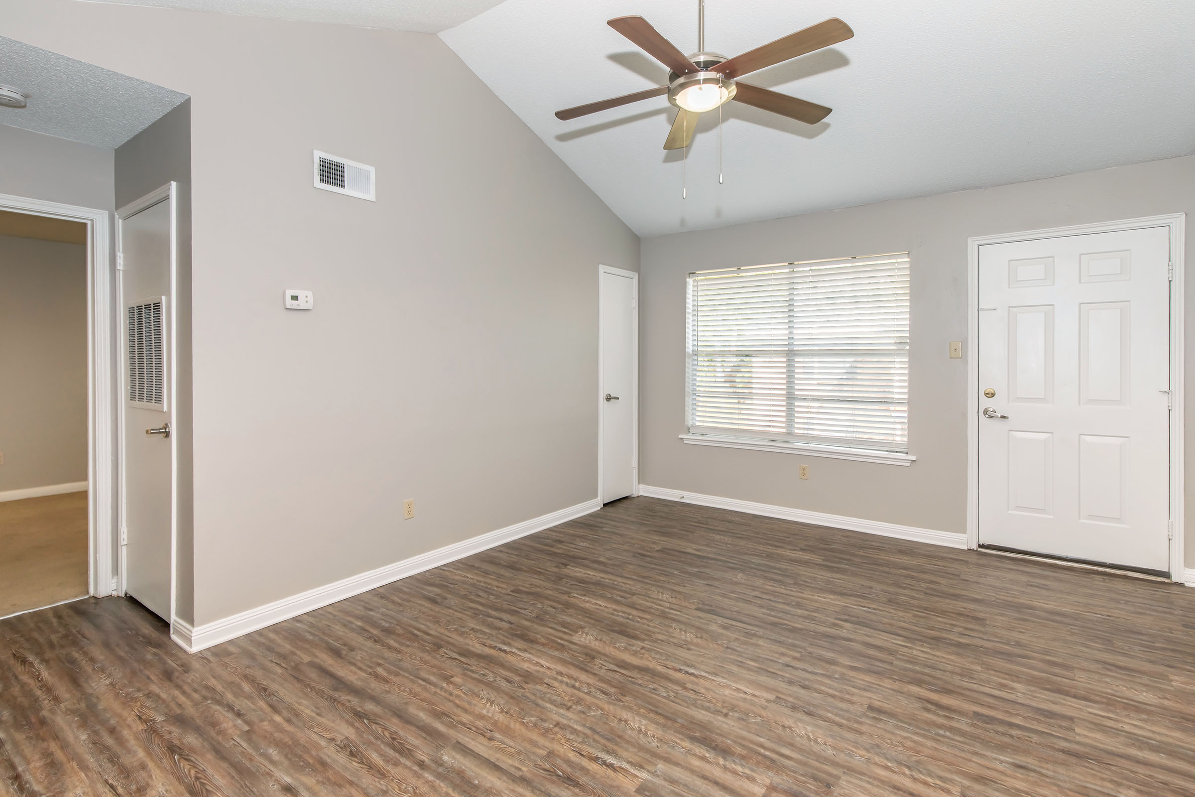 A bright and spacious living room featuring a vaulted ceiling with a ceiling fan, light-colored walls, and laminate flooring. There are two doorways leading to other rooms, a window with blinds letting in natural light, and a white front door. The atmosphere is warm and inviting.