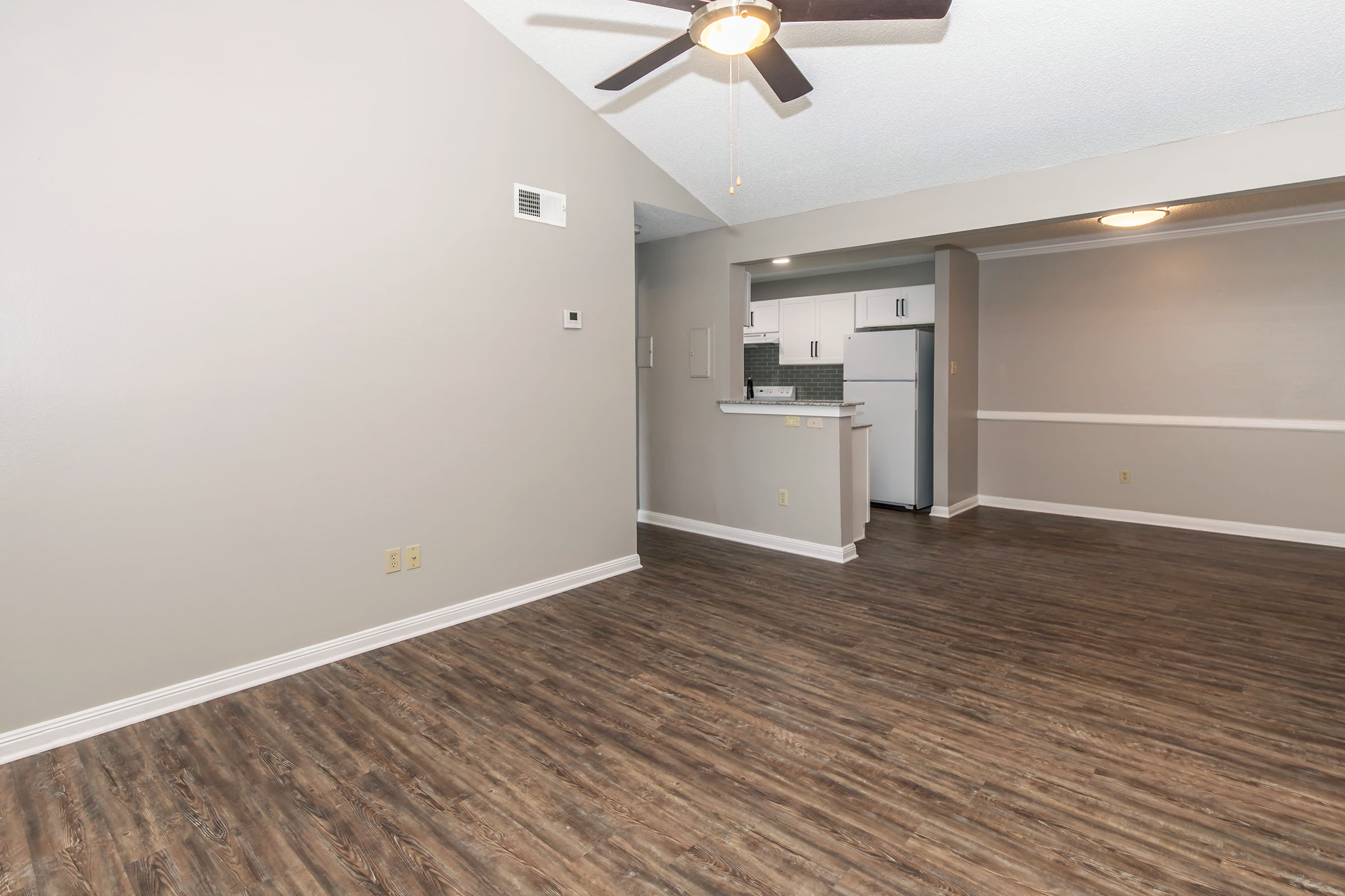 A spacious living area with a ceiling fan, featuring light gray walls and wooden flooring. In the background, a small kitchen is visible with white cabinets and appliances. The room is well-lit with natural light and has a clean, modern aesthetic.
