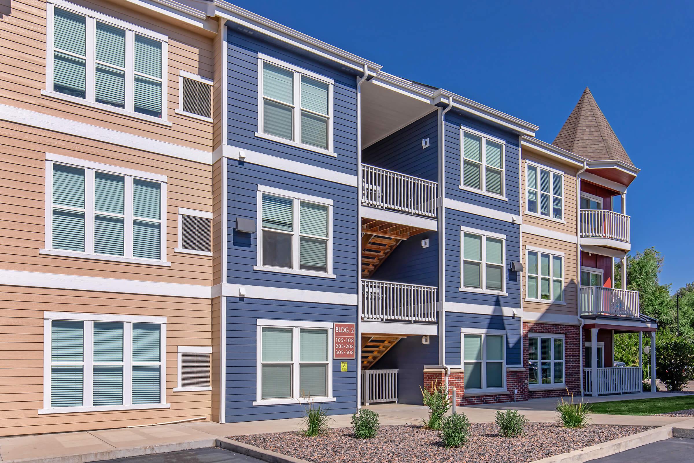 Three-story apartment building featuring a mix of pastel colors, including peach, blue, and red. Each floor has multiple large windows and balconies, with exterior stairs located on the left side. Decorative elements include brick accents and a peaked roof on one section. Clear blue sky in the background.