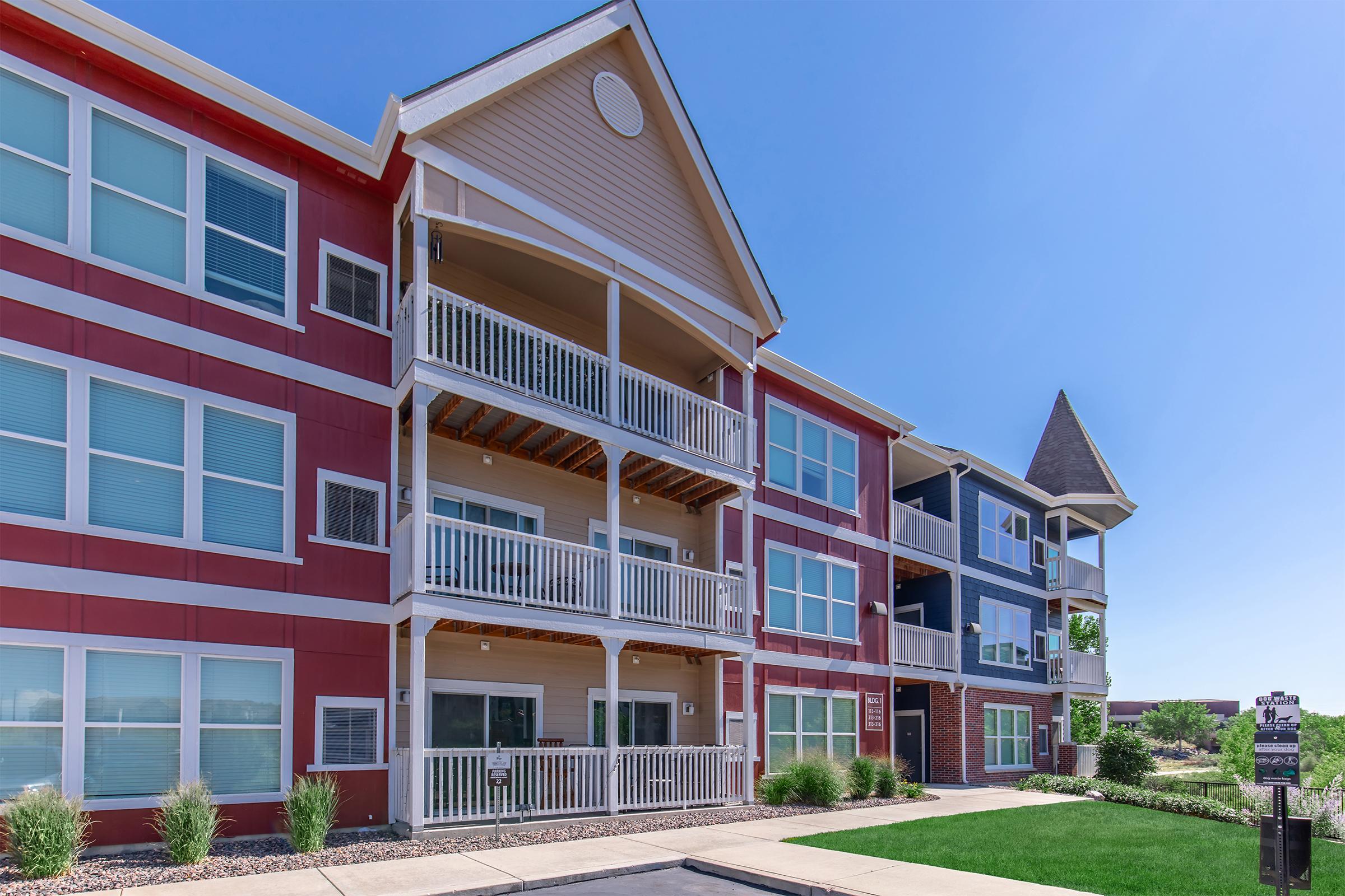 A modern multi-story apartment building featuring a mix of red, blue, and beige siding. The structure has multiple balconies, large windows, and a well-maintained lawn. Clear blue sky provides a bright and inviting atmosphere. The entrance is visible, with pathways leading to the building.