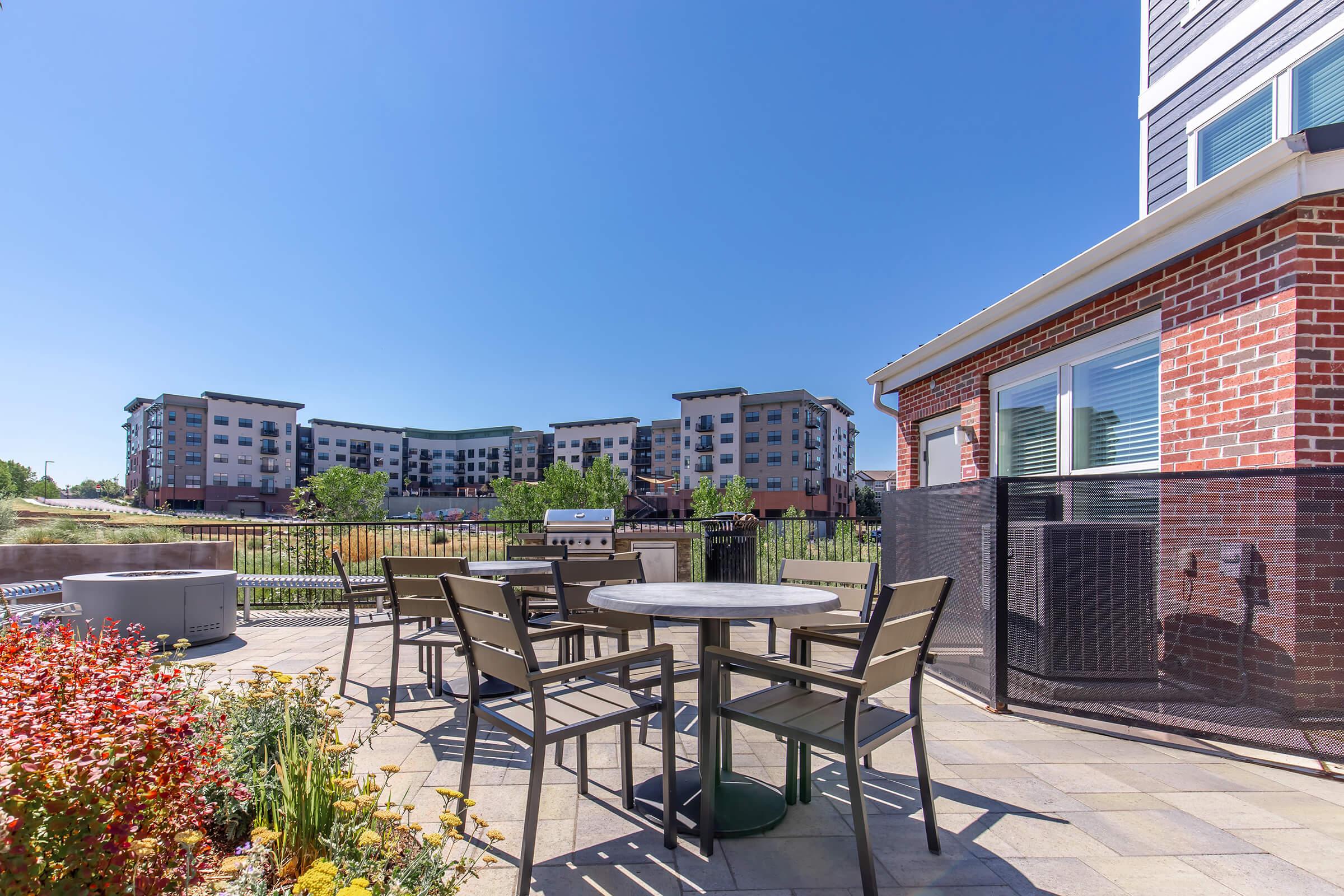 Outdoor seating area with a round table and several chairs, surrounded by colorful flowers. In the background, there are modern apartment buildings under a clear blue sky. A grill is visible nearby, indicating a communal space for residents. Bright and inviting atmosphere.