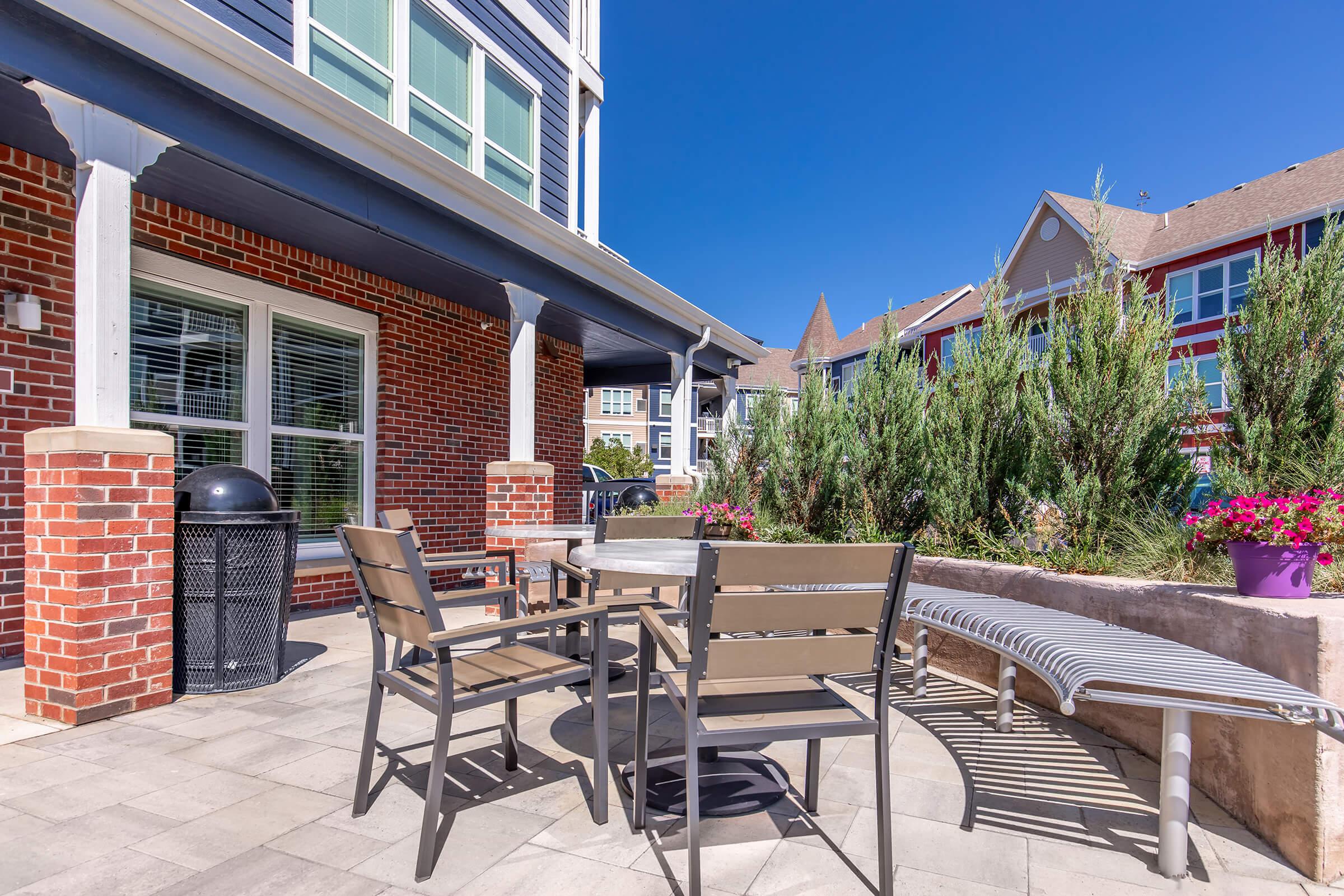 A patio area featuring a round table with chairs, surrounded by greenery and colorful flower pots. There's a trash can in the corner and a bench along the side, with a bright blue sky overhead and nearby buildings visible in the background.
