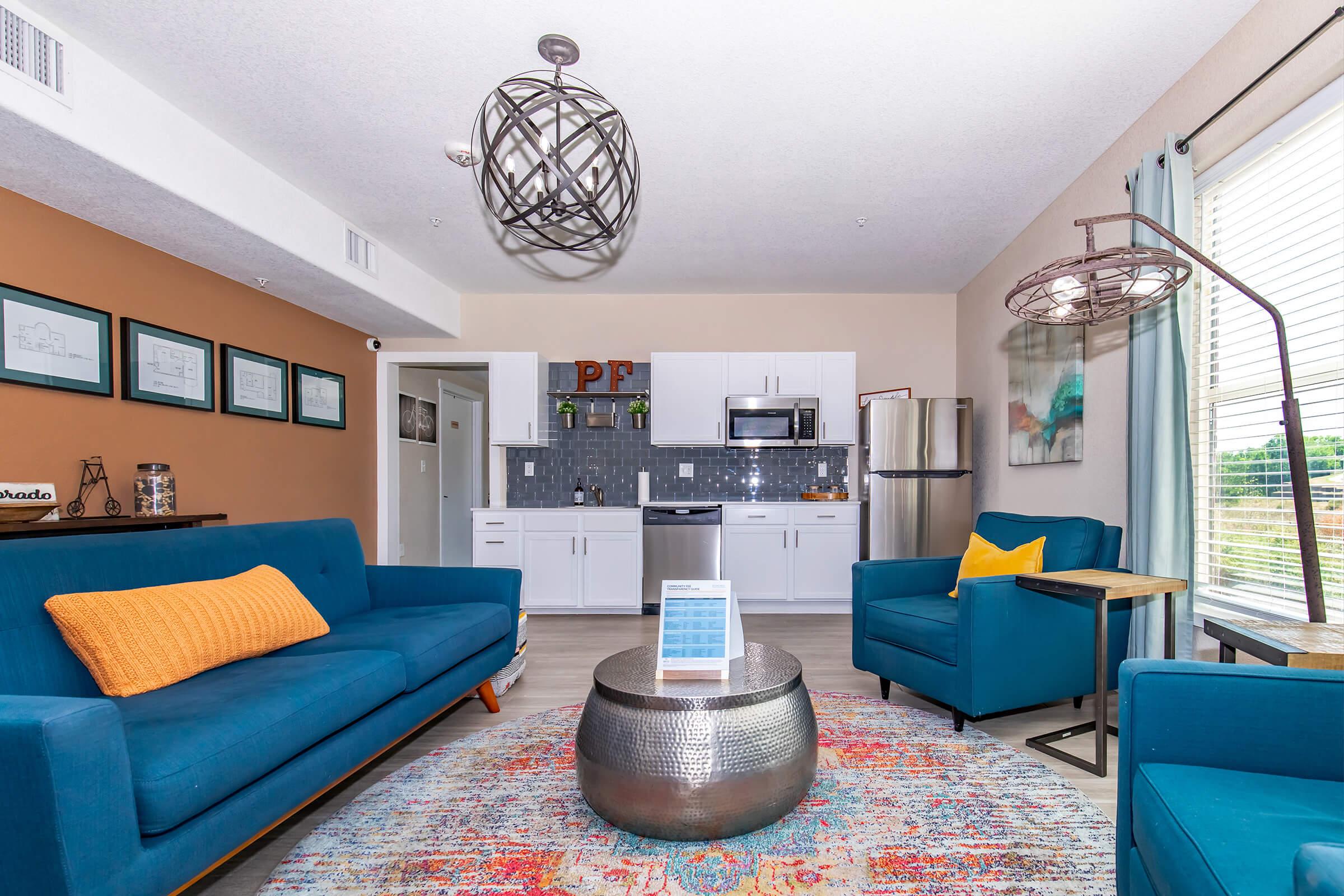 A modern living room featuring two blue sofas with orange cushions, a round metallic coffee table, and a decorative rug. The space has light-colored walls, a kitchen area with stainless steel appliances, and large windows allowing natural light. Stylish light fixtures complement the contemporary decor.