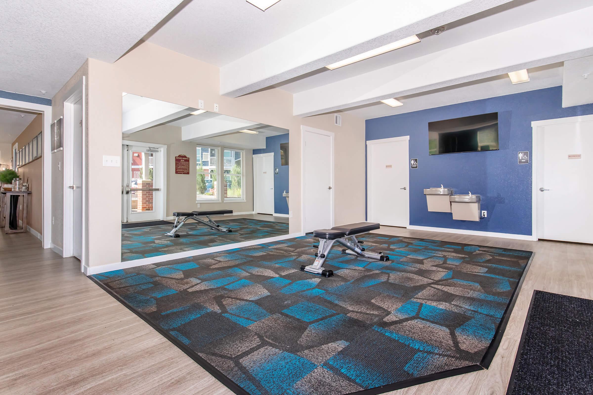 A fitness room featuring a bench press, mirrored wall, and colorful carpet. Large windows provide natural light, revealing outdoor views. There is a water fountain and a wall-mounted TV. The walls are painted in light and dark tones, creating a modern and inviting atmosphere.