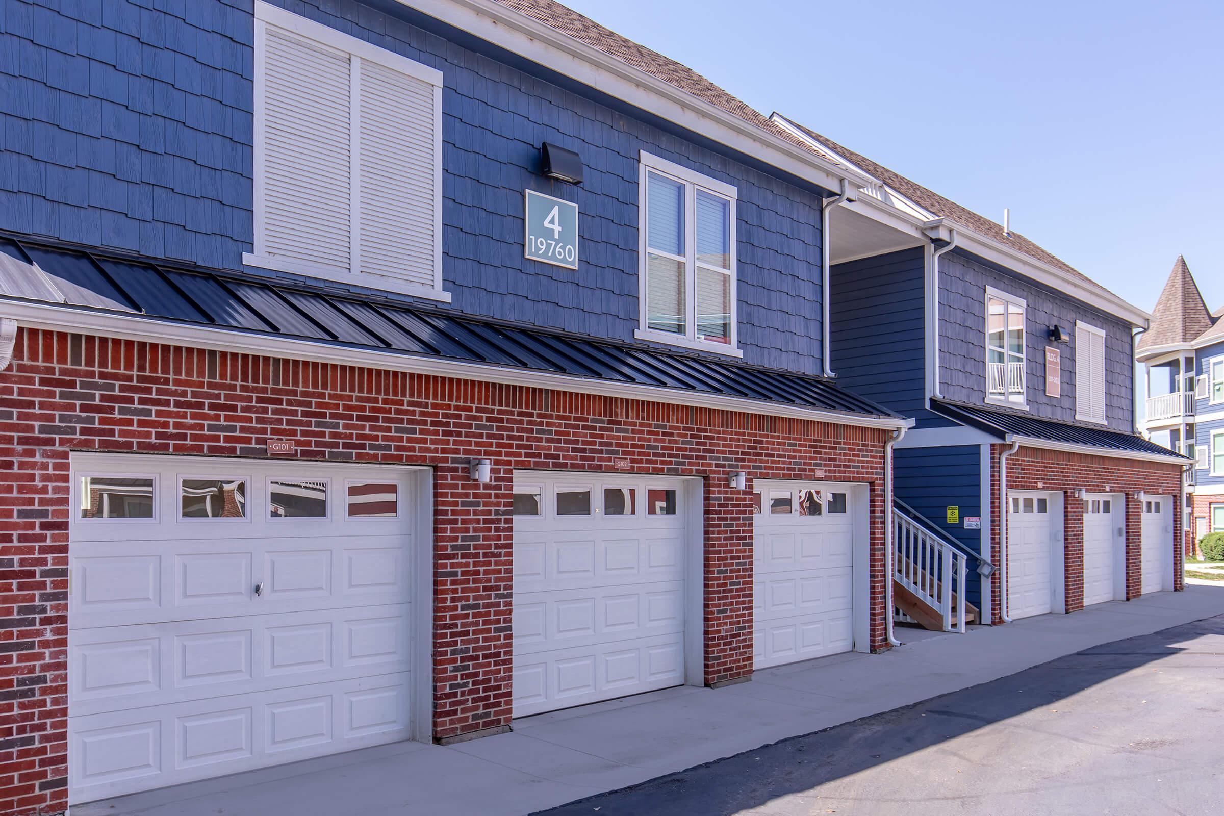 A row of residential garage doors with a blue shingled upper story. The building features a brick facade, large windows, and a visible number sign. A staircase leads to the upper level, and the setting is well-lit with clear blue skies in the background.