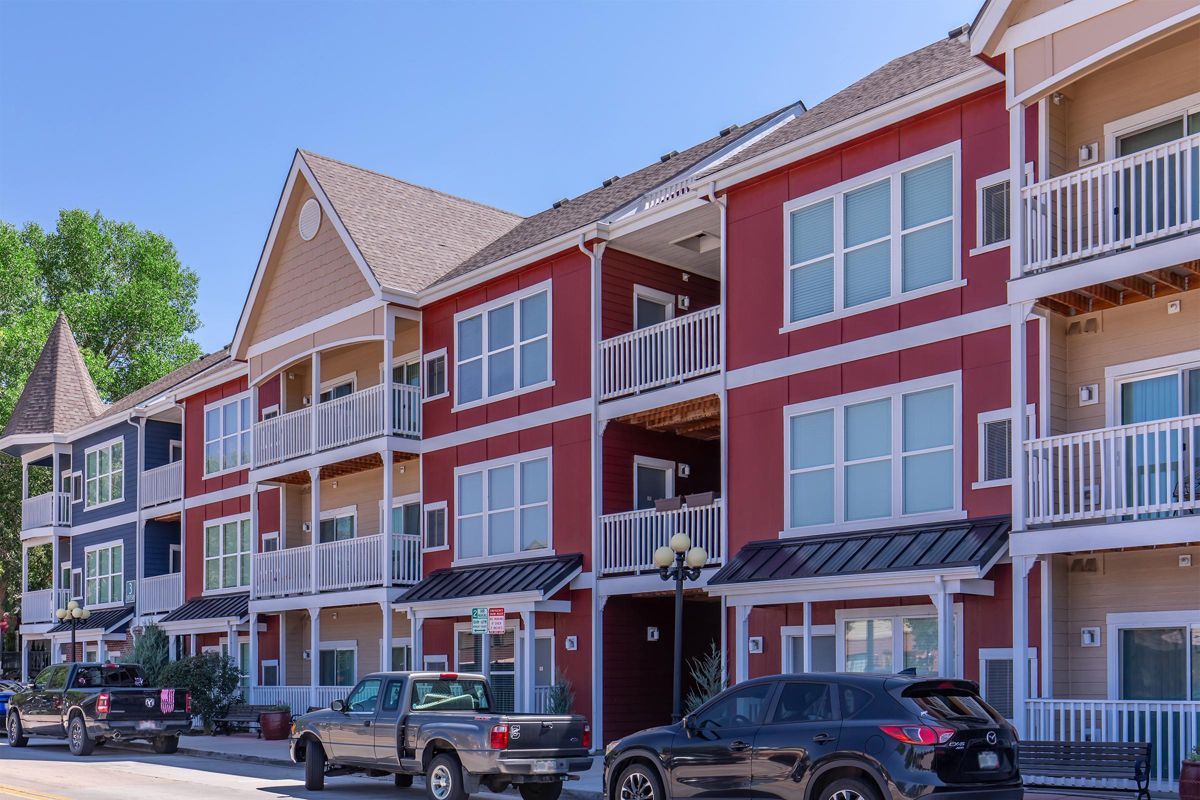 A row of modern apartment buildings featuring a mix of red and beige siding. Each unit has multiple balconies with white railings. Several parked vehicles line the street in front of the buildings, and clear blue skies overhead complement the vibrant scene.
