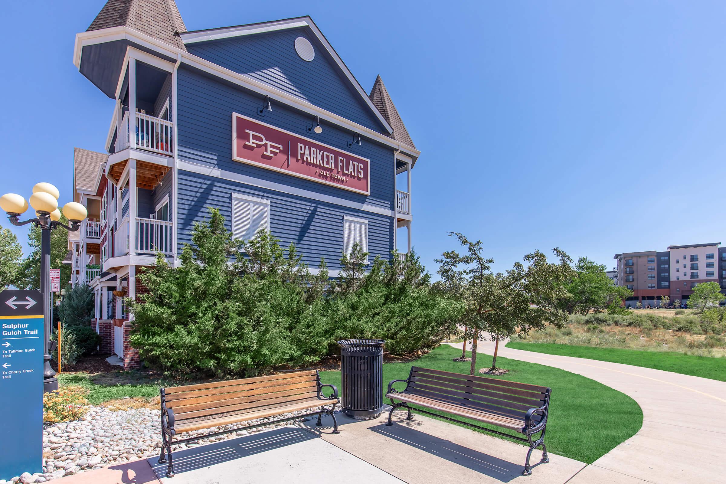 Multi-story blue building with a prominent sign reading "Parker Flats." Surrounding greenery and two benches are in the foreground, along with a trash can. A pathway leads away from the building, and additional buildings can be seen in the background under a clear blue sky.