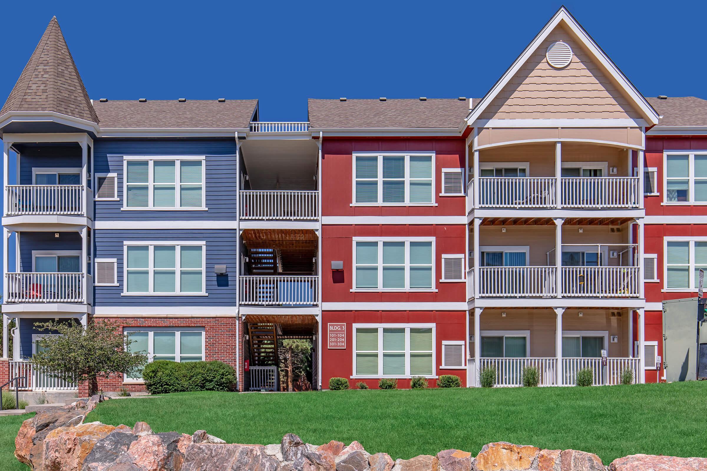 A colorful apartment building featuring blue and red exteriors with balconies. The structure has multiple floors, large windows, and is set against a bright blue sky. A well-maintained lawn with rocks in the foreground adds to the appealing landscape.