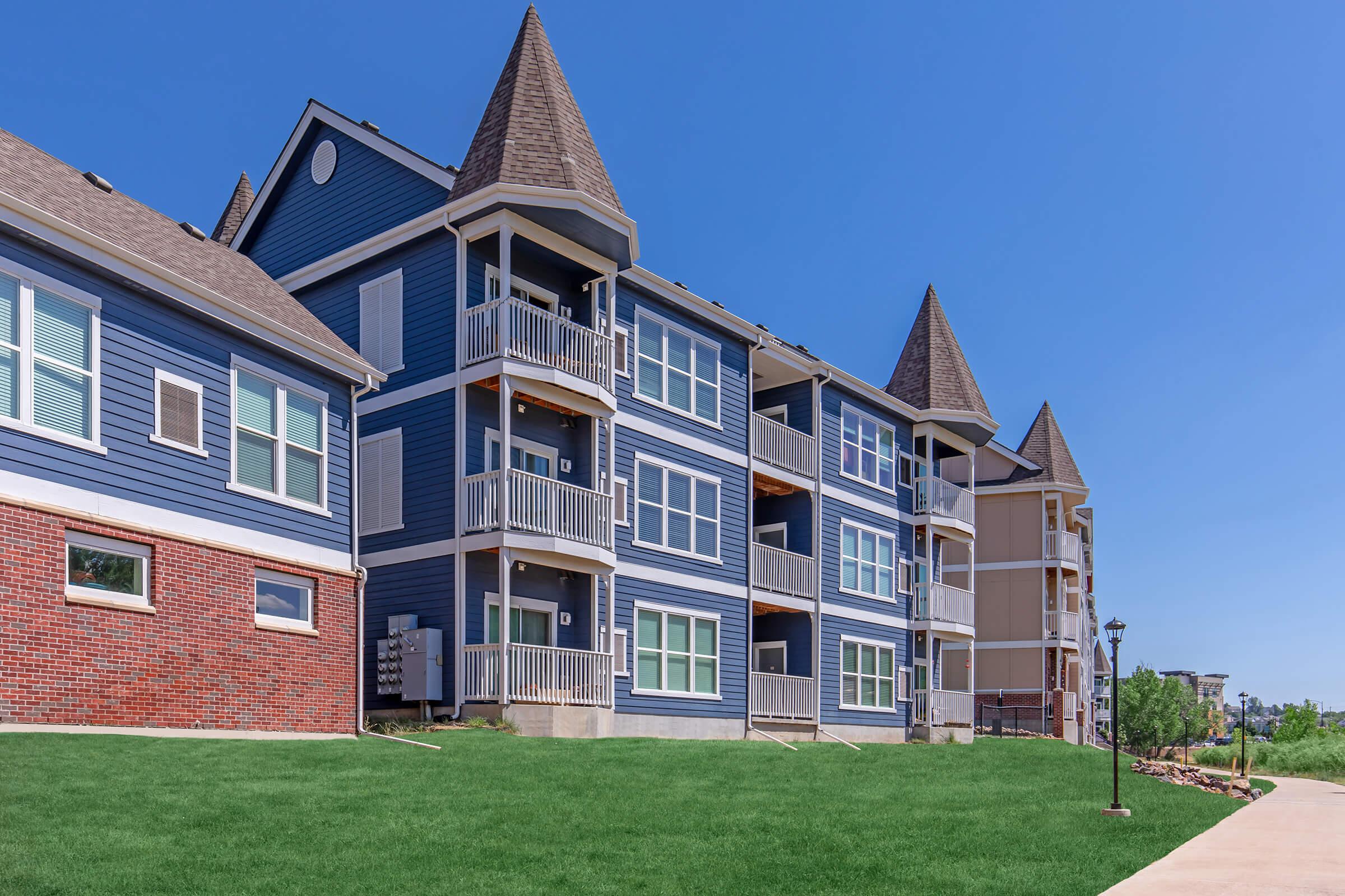 A row of modern multi-story apartment buildings featuring blue siding and distinctive peaked roofs. The buildings are surrounded by well-maintained green lawn and landscaping, with a clear blue sky above and a walking path in the foreground.